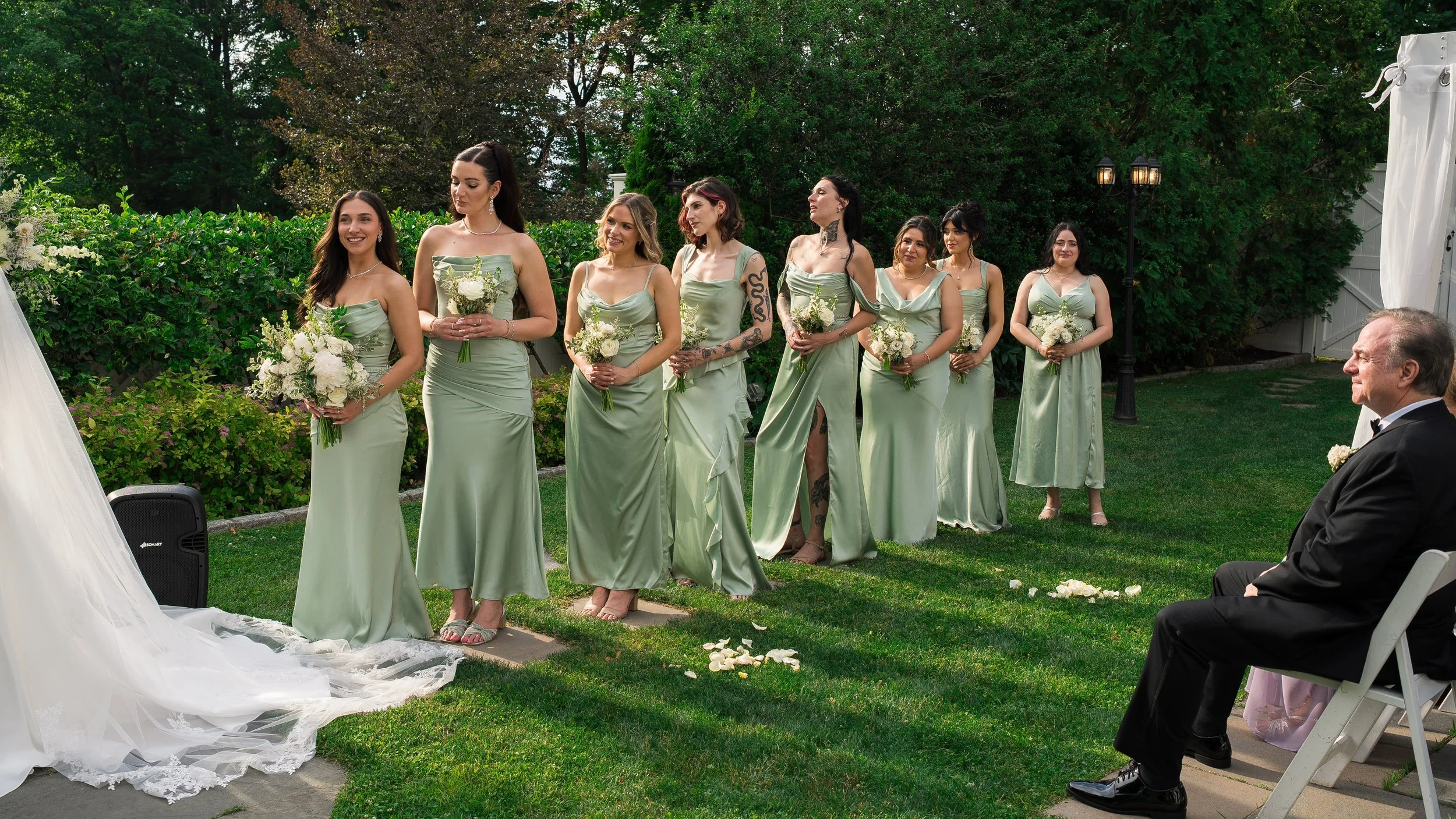 A group of eight women in matching light green dresses stand outdoors during a wedding ceremony, holding white floral bouquets.