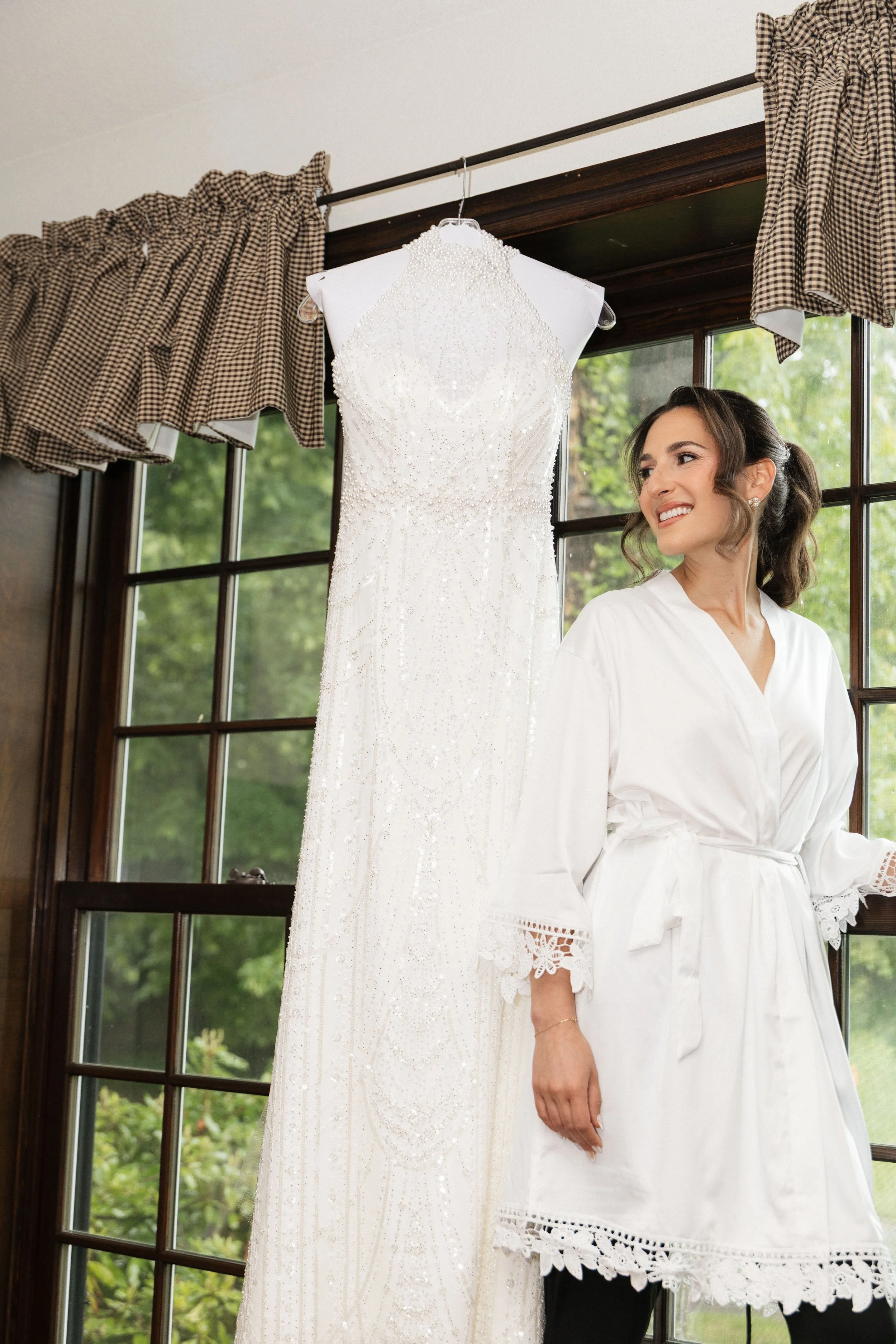 A woman with brown hair in a ponytail smiling while looking at a wedding dress hanging on a hanger near a window with checkered curtains and green trees outside.