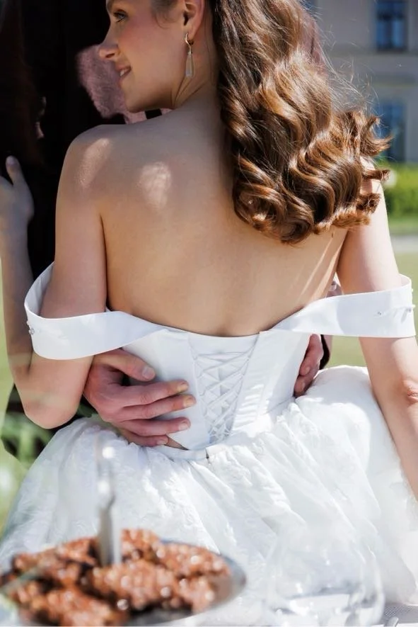 A woman in a white off-the-shoulder dress, smiling and being held by someone, at what appears to be a celebration or wedding, with food in the foreground.