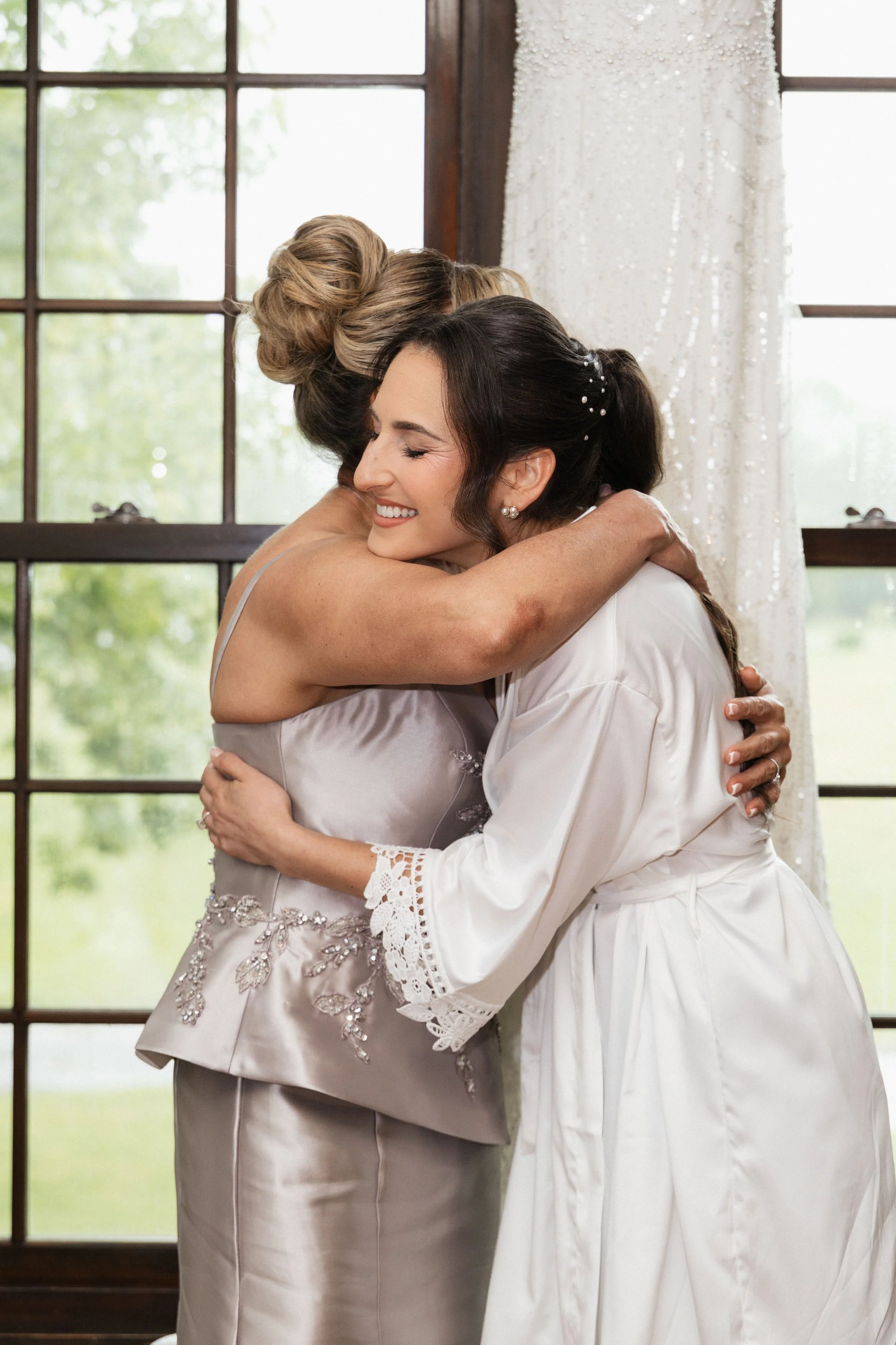 Two women hugging warmly in front of a window with natural light, one in a wedding dress and the other in a satin dress with embroidery.