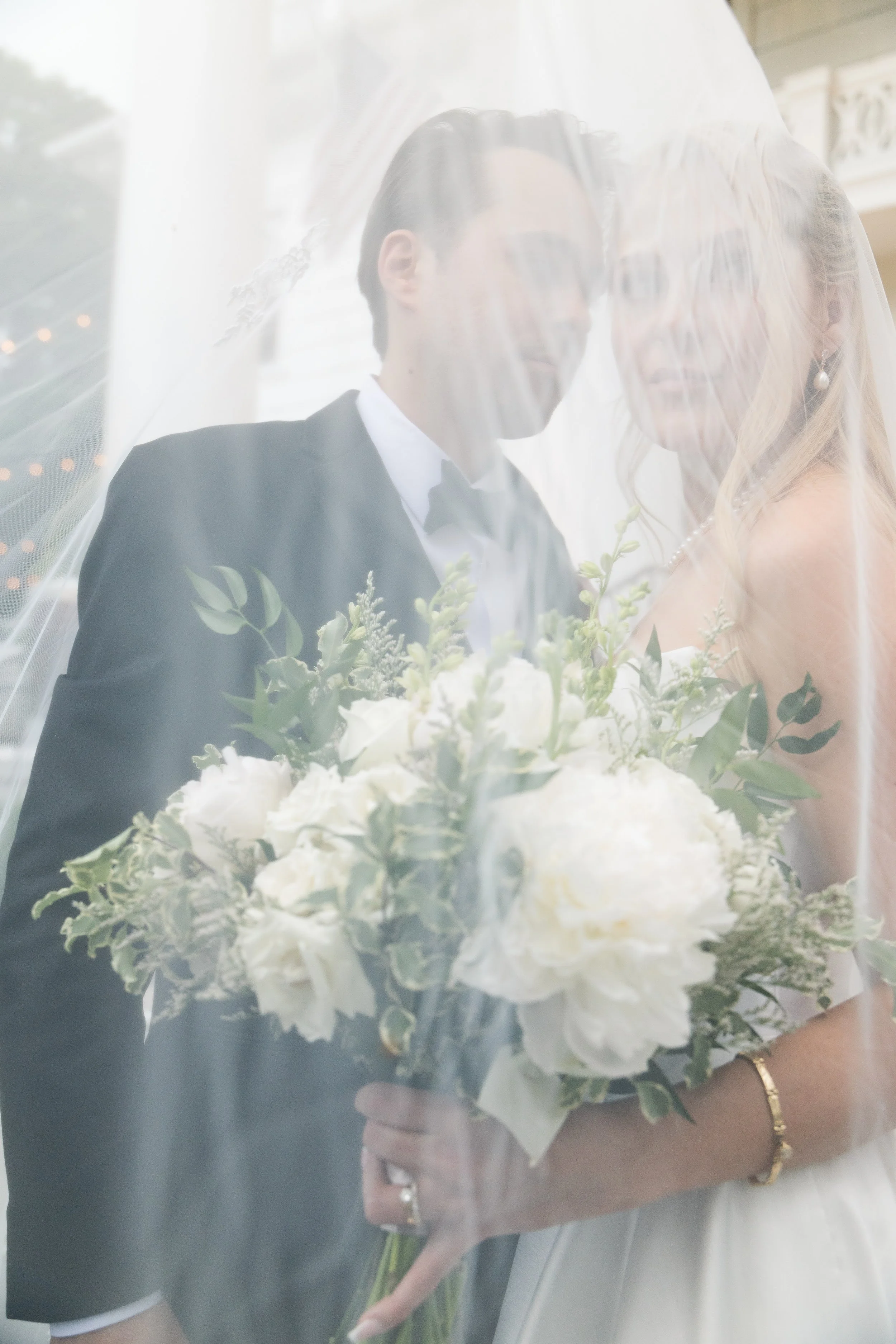 A bride and groom are standing close together, partially obscured by a sheer fabric veil. The bride is holding a bouquet of white flowers with green foliage. The groom is wearing a black tuxedo, and the bride is wearing a strapless wedding dress with