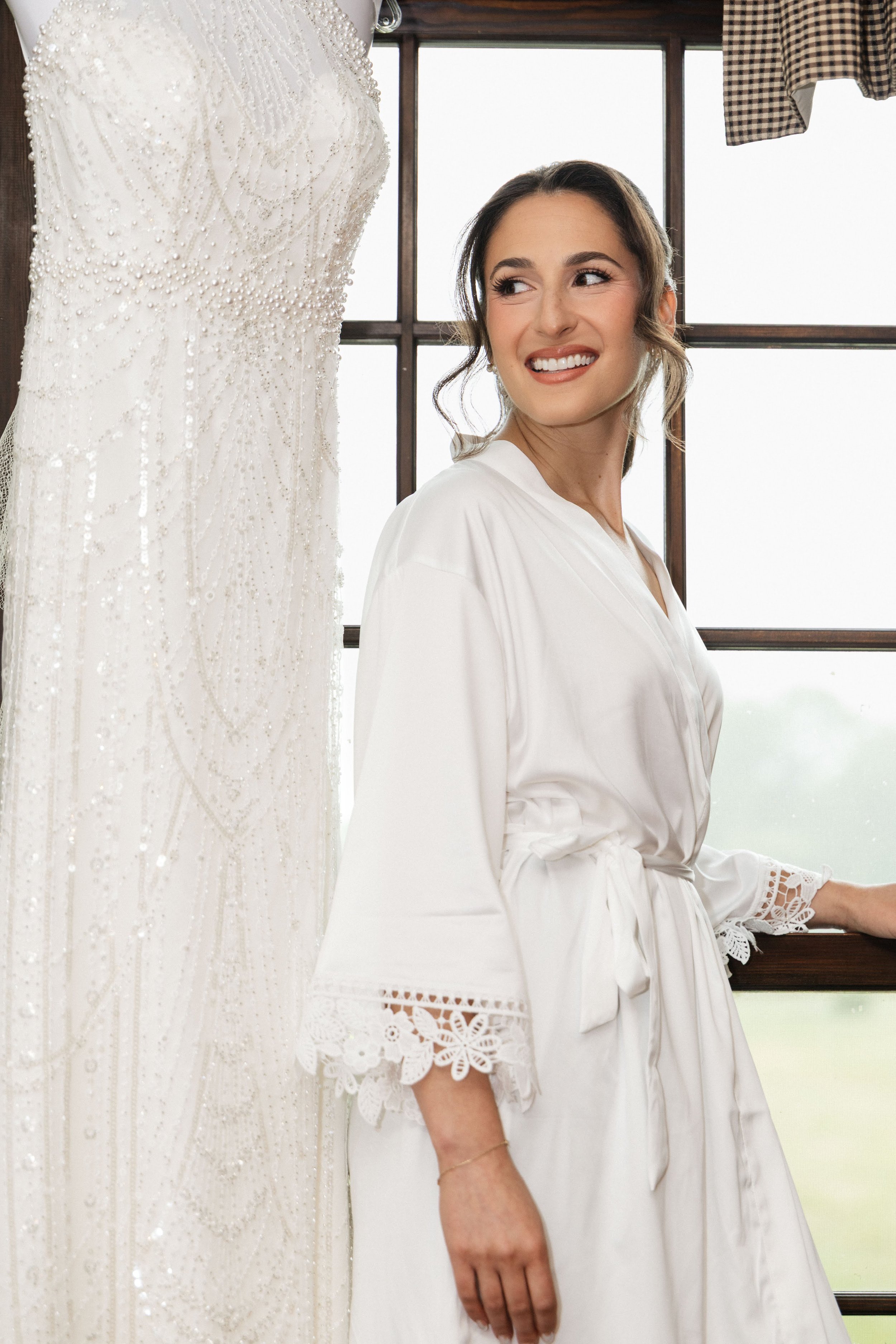 A woman in a white robe smiling while looking to the side near a wedding dress with beaded embroidery hanging on a mannequin, in front of a window with wooden frames.