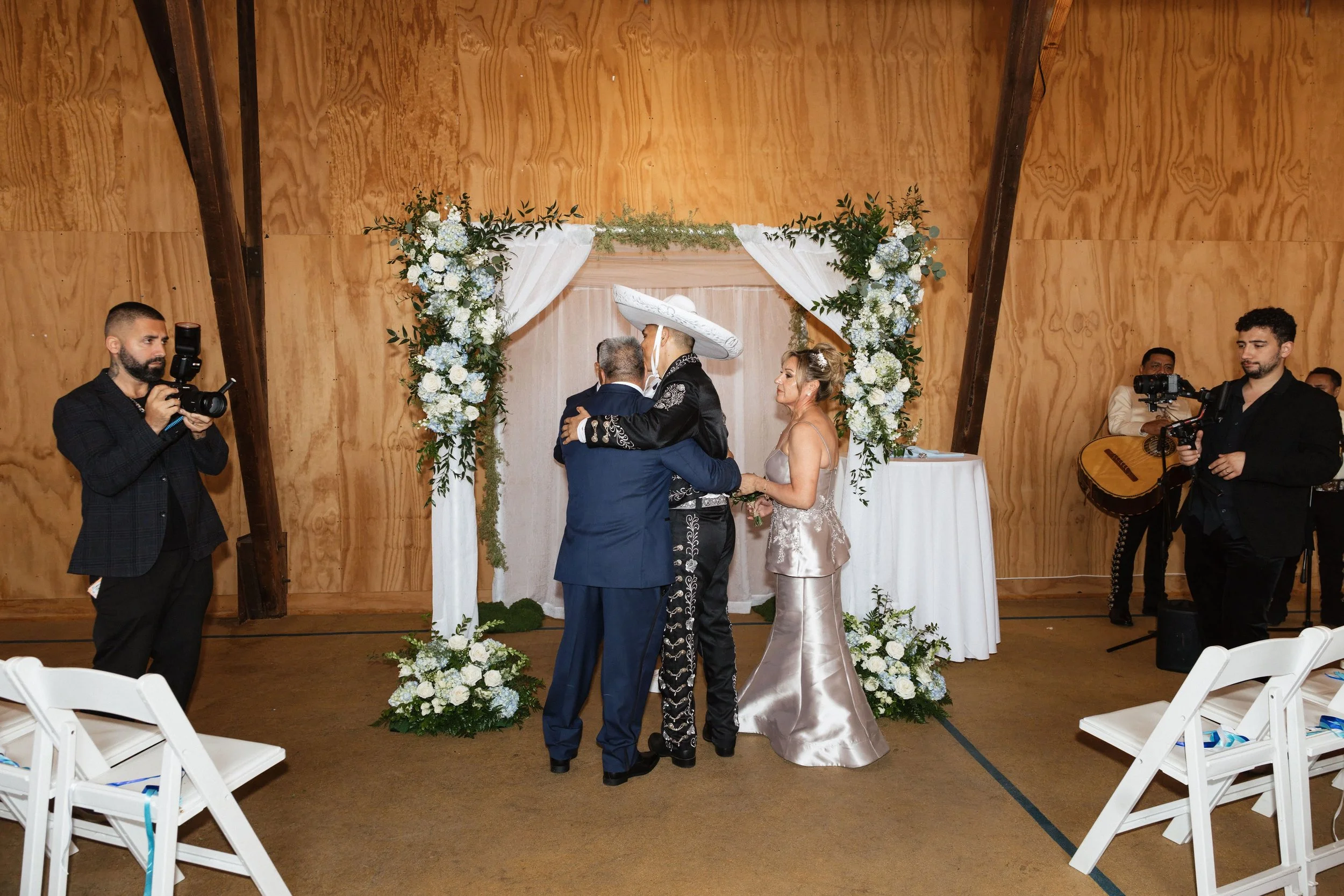 People attending a wedding ceremony under a floral arch with wooden walls in the background.