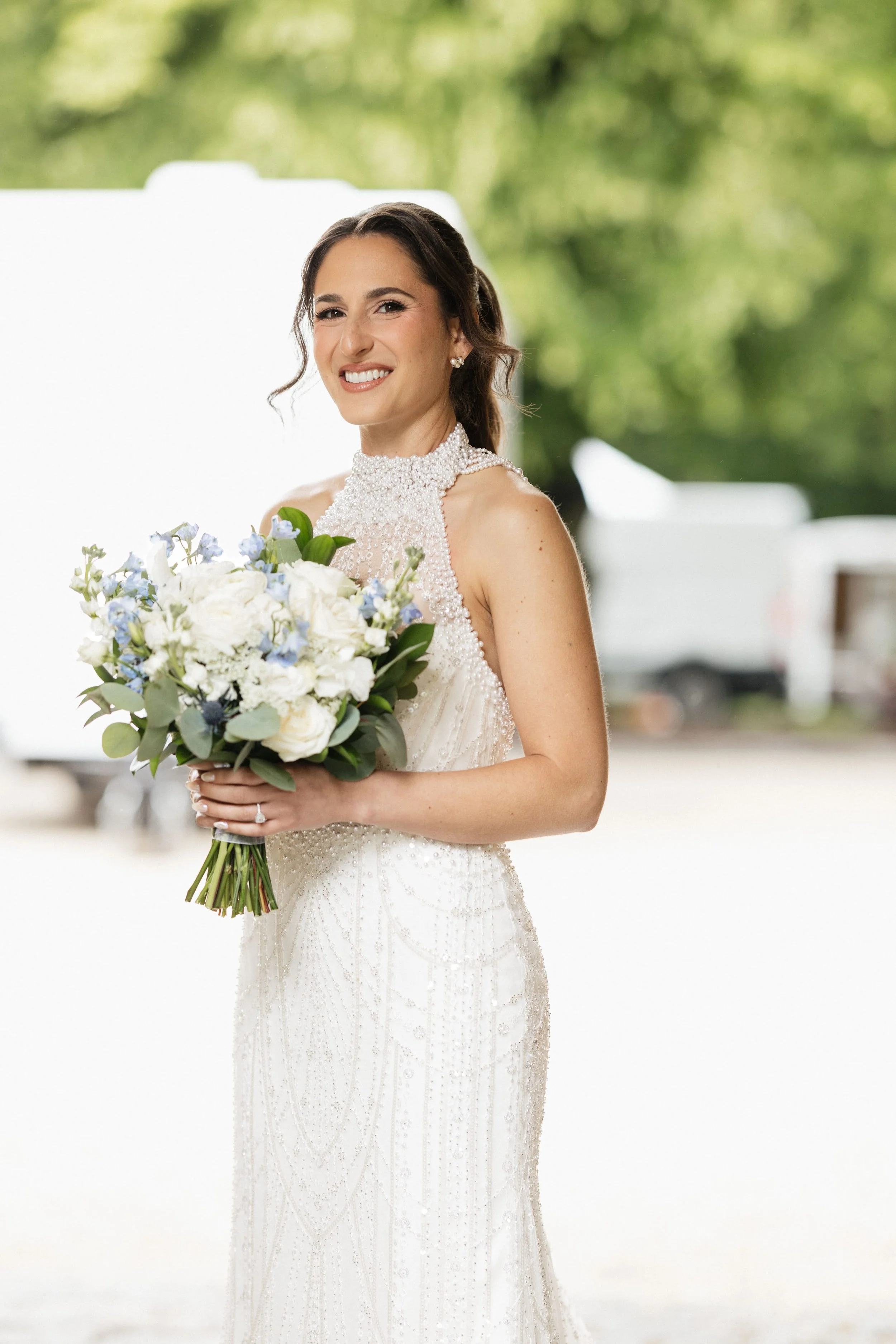 A bride in a detailed white wedding gown holding a bouquet of white and light purple flowers, smiling outdoors with green trees and a white tent in the background.