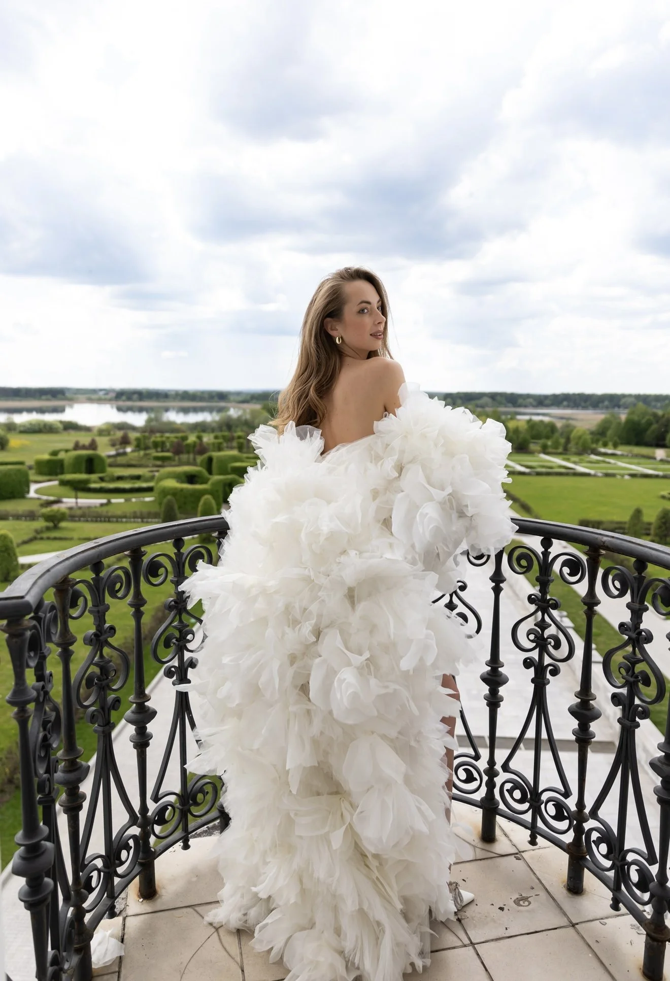 A woman in a white, ruffled gown stands on a balcony with a decorative black railing, overlooking a lush green landscape with trees, fields, and water under a cloudy sky.