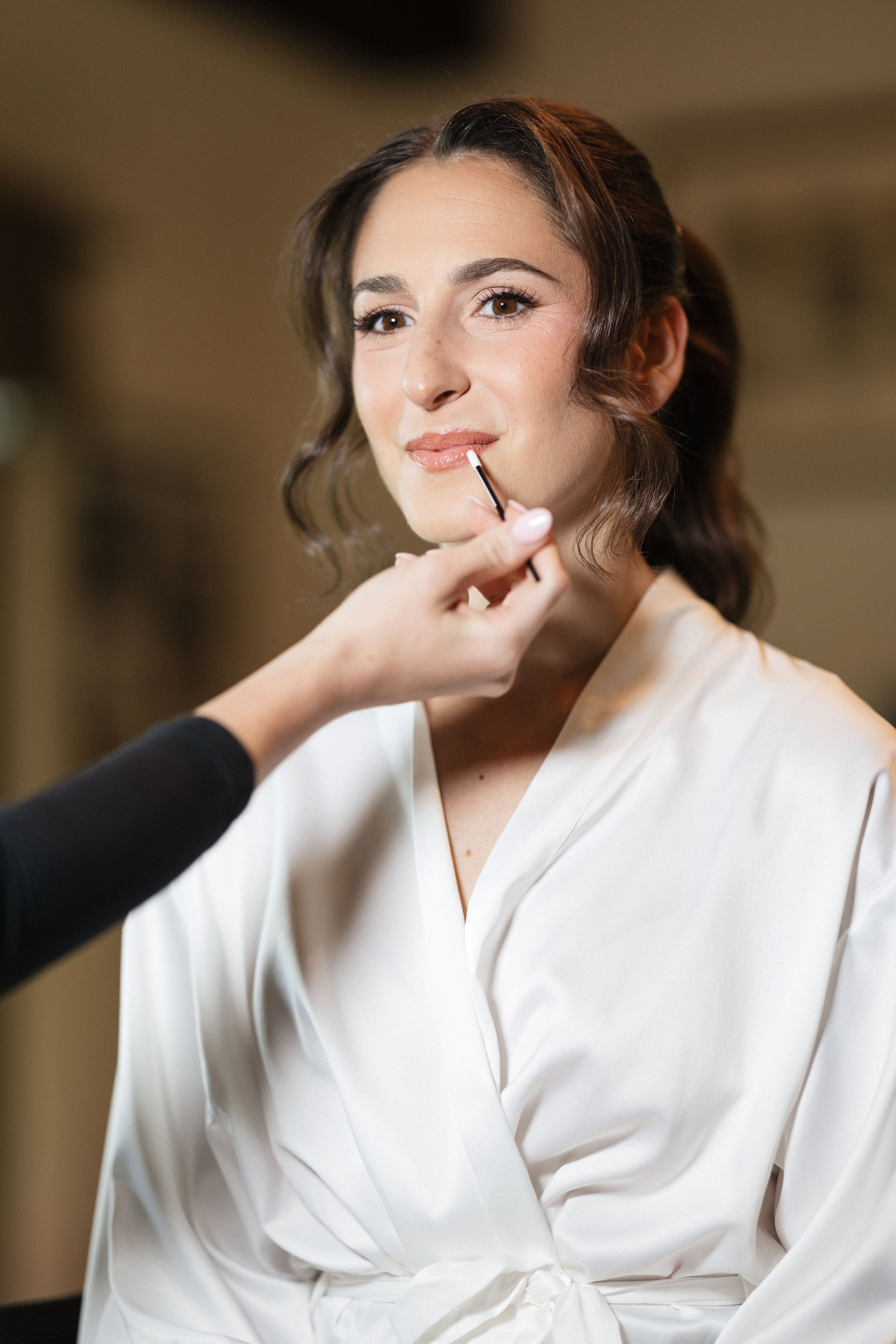 A woman with shoulder-length wavy brown hair is having her lips painted with makeup by a person whose hand is visible. The woman is wearing a white satin robe and is looking slightly off-camera with a gentle smile.