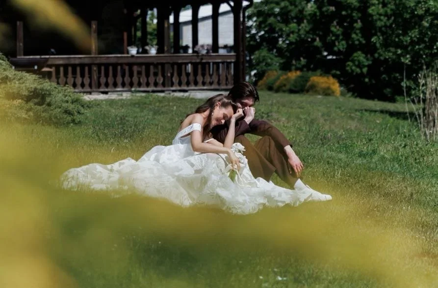 A bride and groom sitting on grass in a backyard, sharing a joyful moment, with a house and garden in the background.
