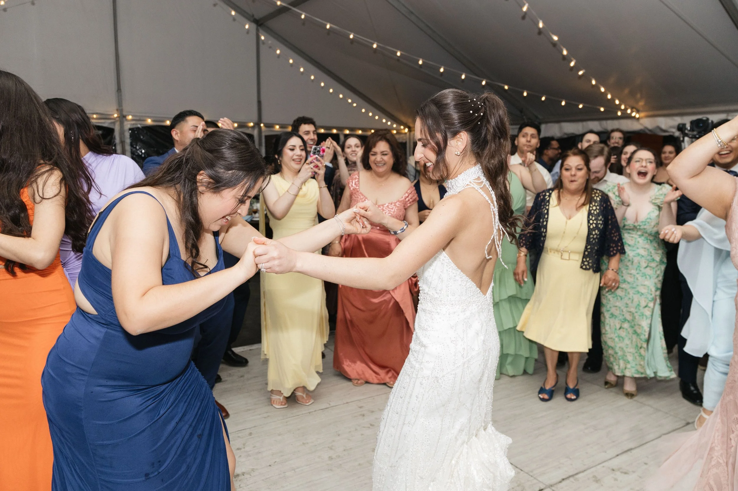 A wedding reception with a bride in a white wedding dress dancing with a guest in a blue dress. Multiple guests near them are smiling, clapping, and taking photos under string lights in an indoor tent.