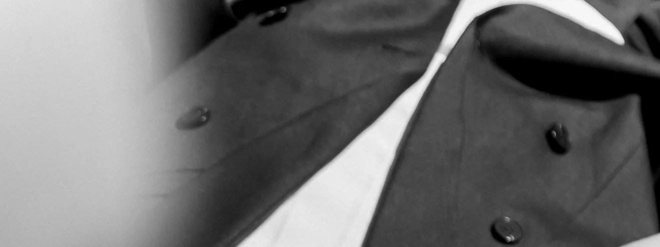 A close-up of a person wearing a dark suit jacket with white accents and black buttons.
