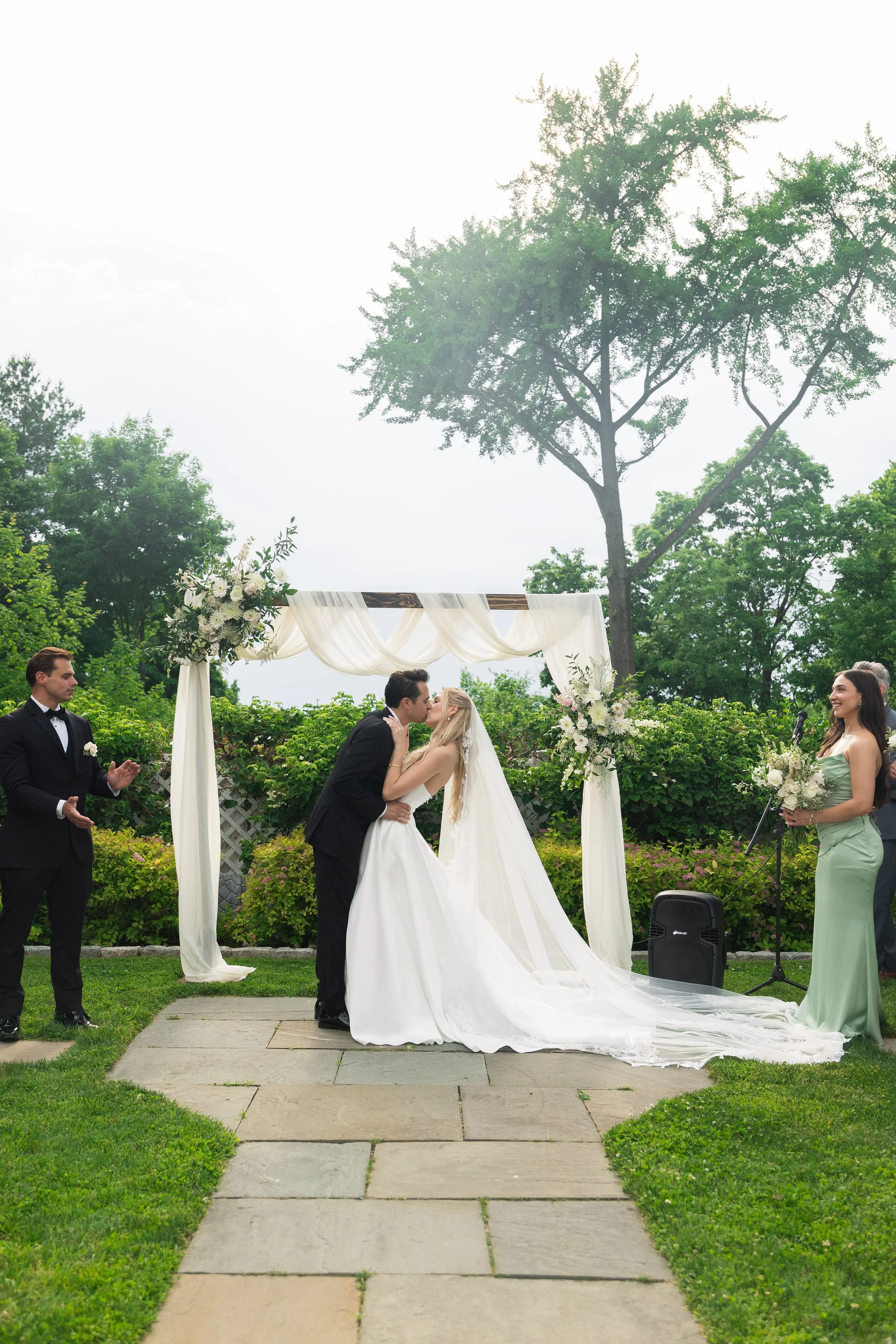 A couple getting married outdoors under a decorated arch, surrounded by greenery, with officiant and guests present.