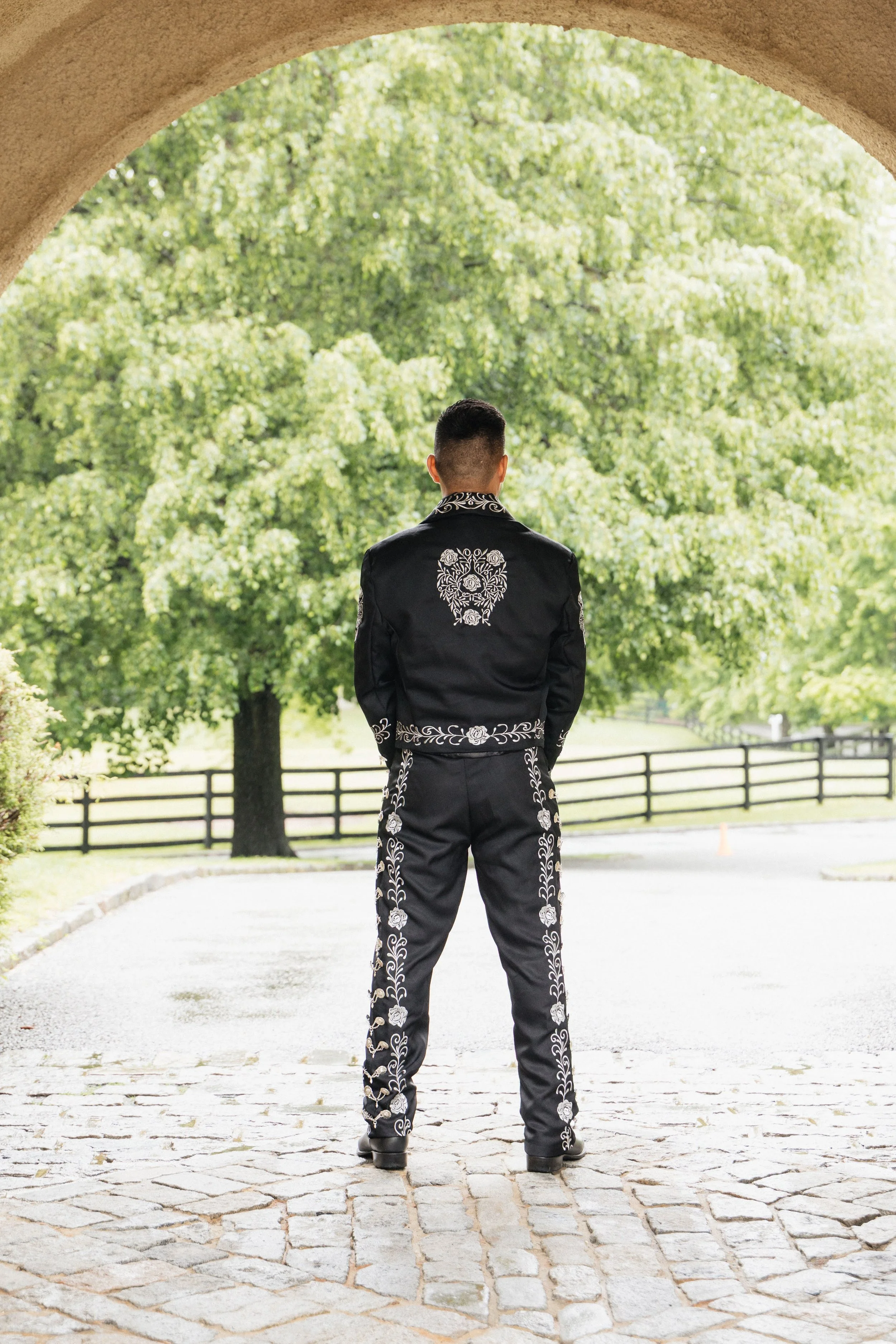 Man standing with his back to the camera, wearing a black embroidered Mariachi suit, under a stone archway with a park and trees in the background.