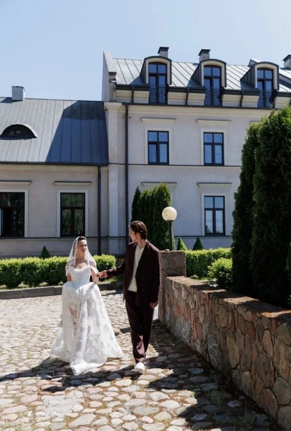 Bride and groom walking hand in hand outside a large, elegant building with dormer windows and a stone wall.