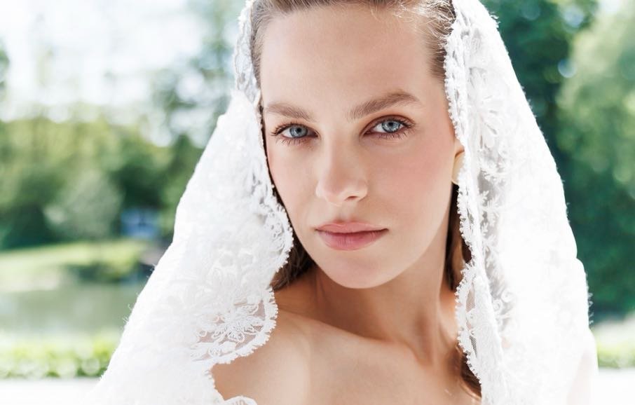 Close-up of a woman with blue eyes and light makeup, wearing a white lace veil outdoors with green trees in the background.