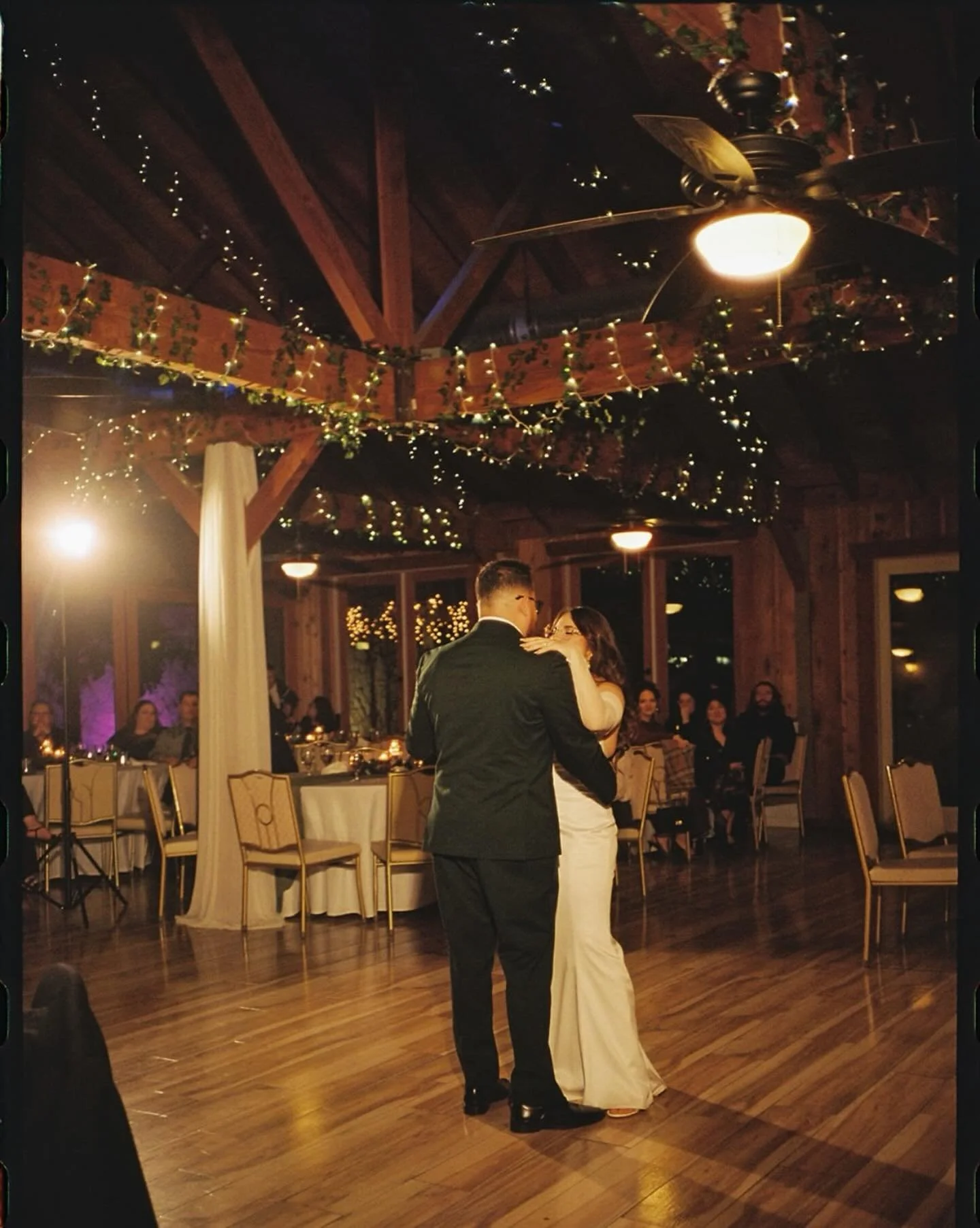 A couple sharing a dance at a wedding reception in a rustic venue with string lights and decorations. The man is in a black suit, and the woman is in a white dress. Guests sit at tables watching them.