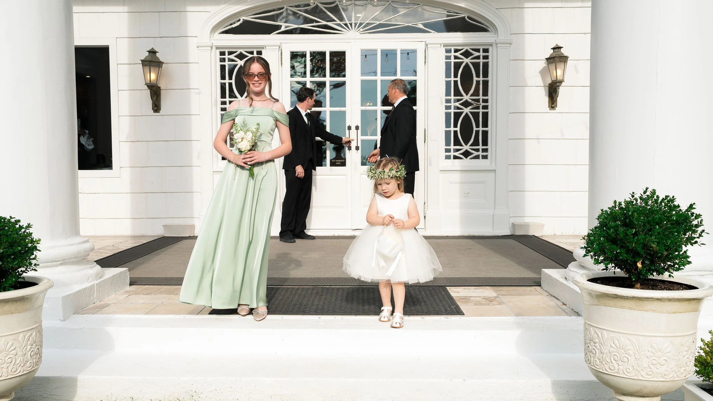 A woman in a light green dress holding a bouquet of flowers, a young girl in a white dress with a flower crown, and three men in black suits standing on the steps of a white building with glass doors, lantern-style lights, and potted plants.