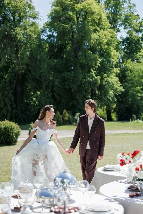 A bride and groom holding hands outdoors at a wedding reception, with tables set with white tablecloths, glassware, and flower arrangements in a lush green park.