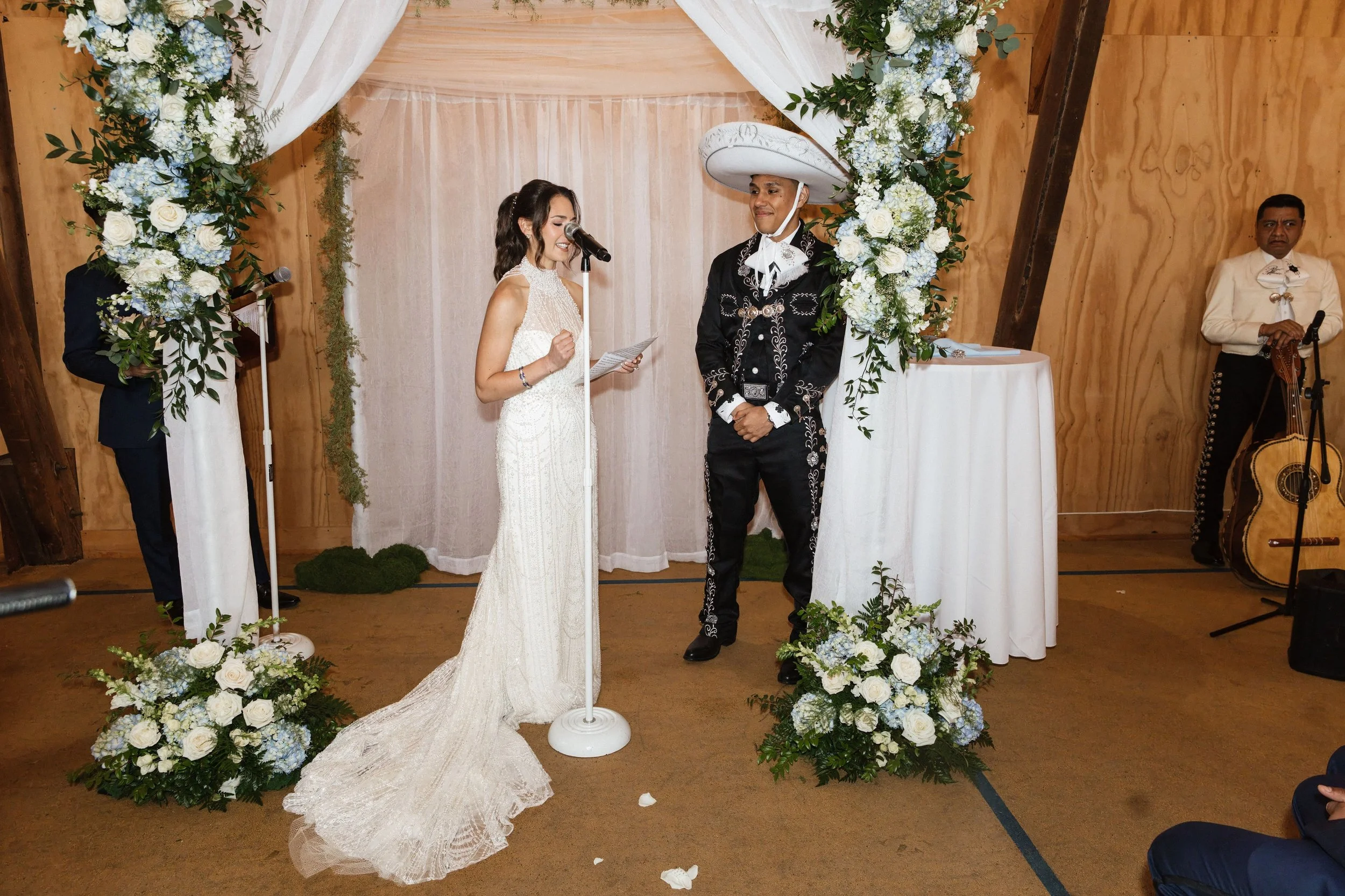 A wedding ceremony with a bride and groom standing under an arch decorated with white and blue flowers, with a woman officiating reading vows, a musician with a guitar on the right, and wooden walls in the background.