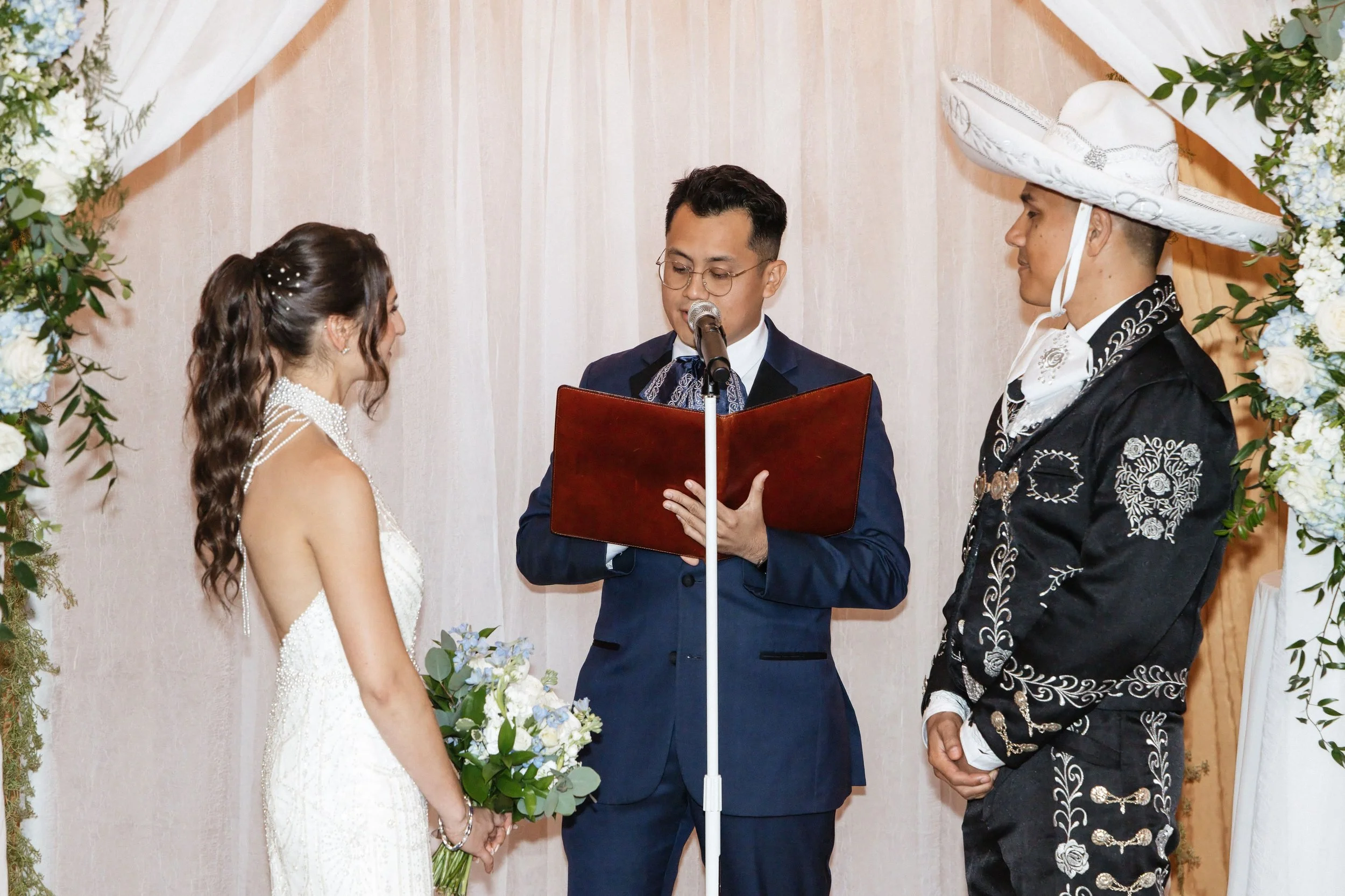 A couple getting married in a ceremony with an officiant reading from a book, in front of floral decorations, with a woman in a wedding dress and a man in a traditional black embroidered suit and large sombrero.