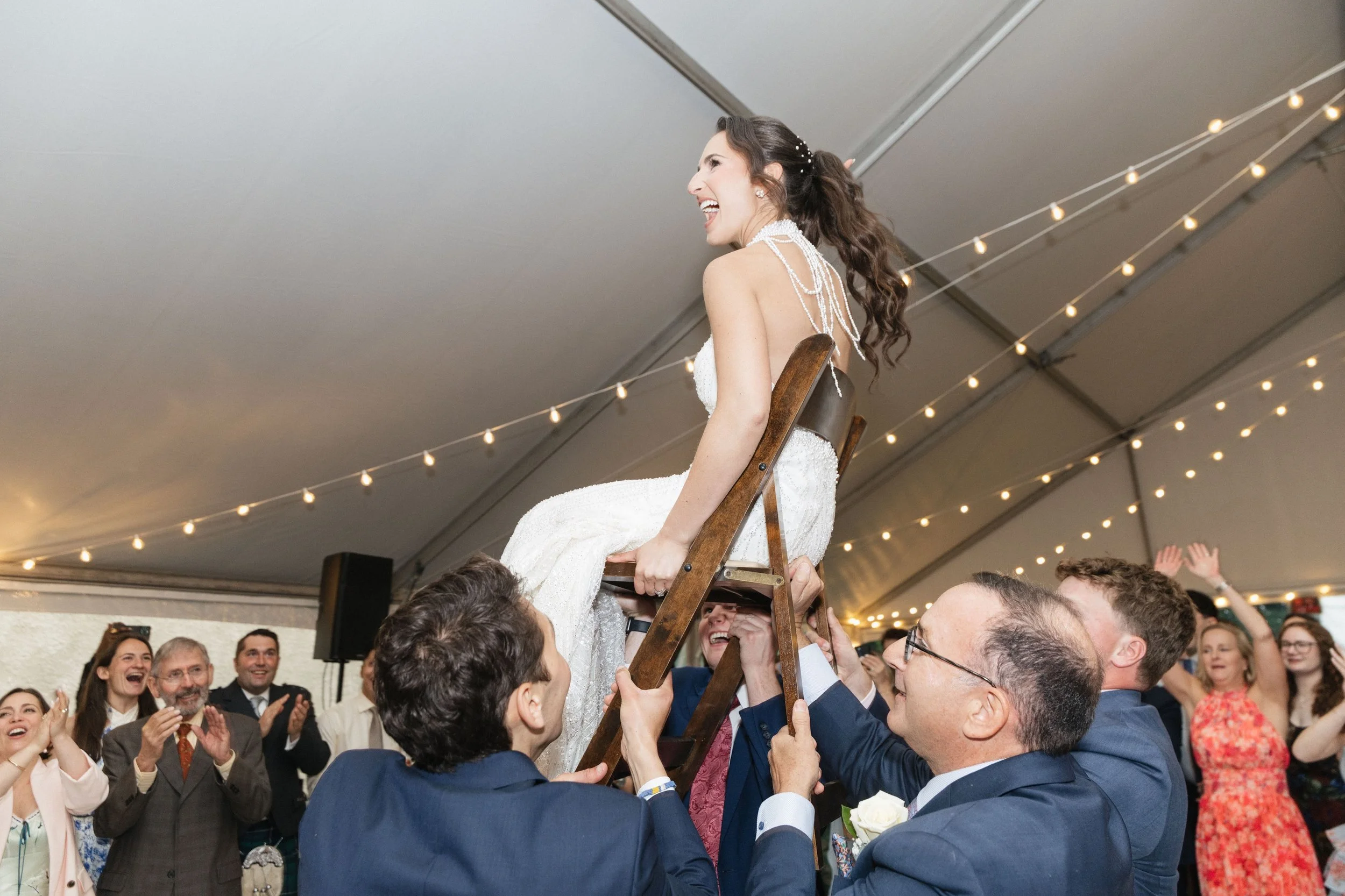 A woman in a wedding dress is being lifted on a chair by several men at a celebration, with people clapping and cheering in the background under a decorated ceiling.
