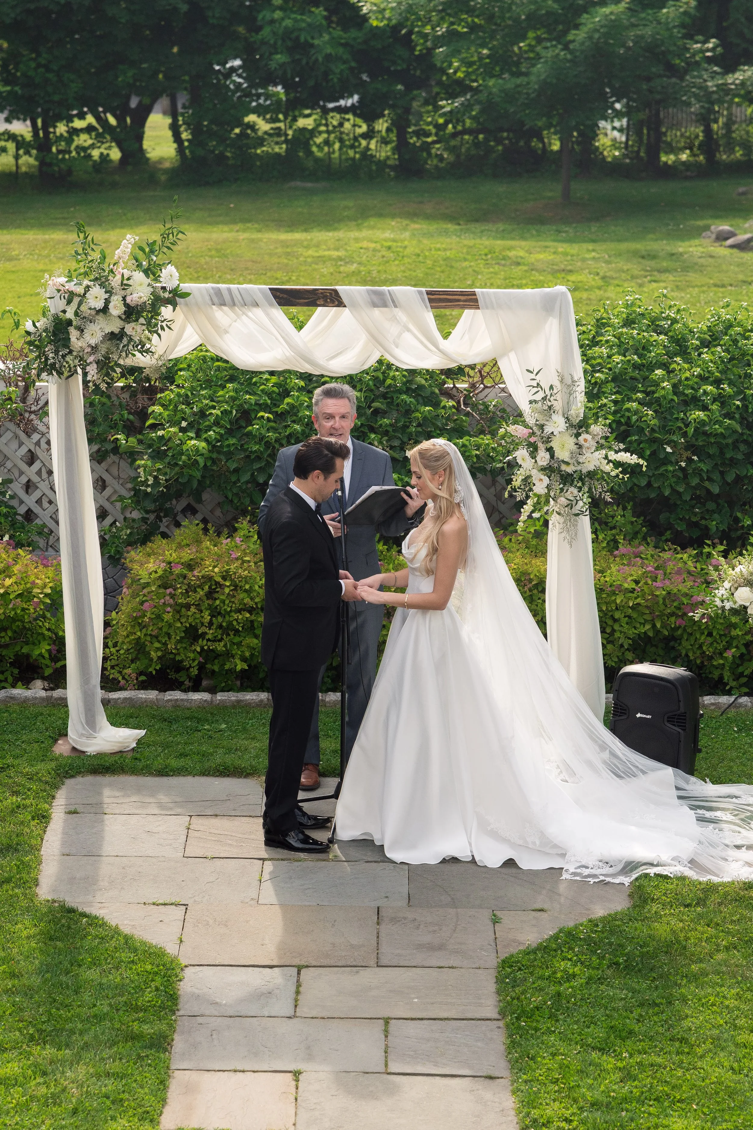 A wedding ceremony takes place outdoors under a white fabric draped arch decorated with white flowers. A couple stands hand-in-hand with an officiant in front of the arch, surrounded by green bushes and trees, on a paved path in a garden.