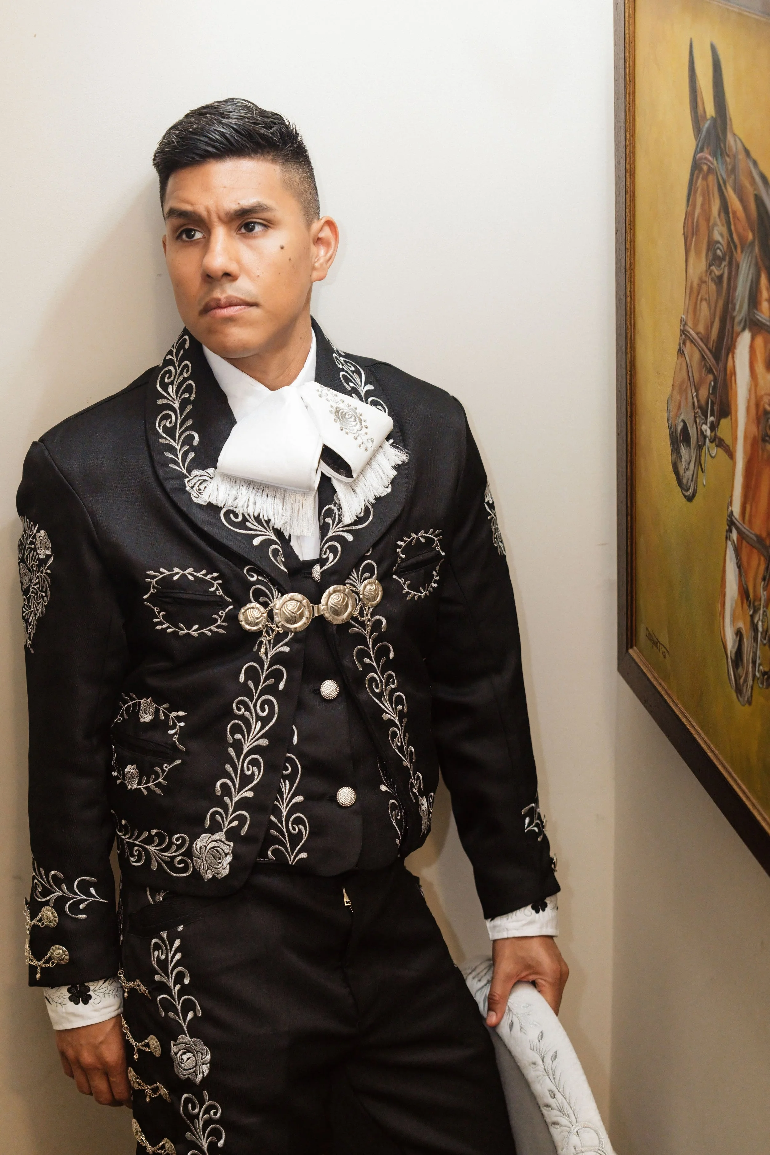 A man dressed in a traditional charro suit, standing against a light-colored wall and looking to his left. There's a painting of a horse on the wall to his right.