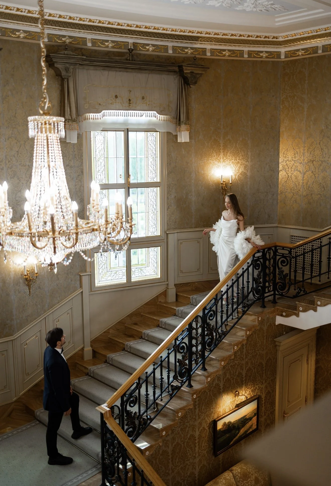 A woman in a white dress standing on a staircase in a luxurious room with ornate gold wallpaper, a chandelier, and decorative window curtains. A man in a suit is standing at the bottom of the stairs looking up at her.