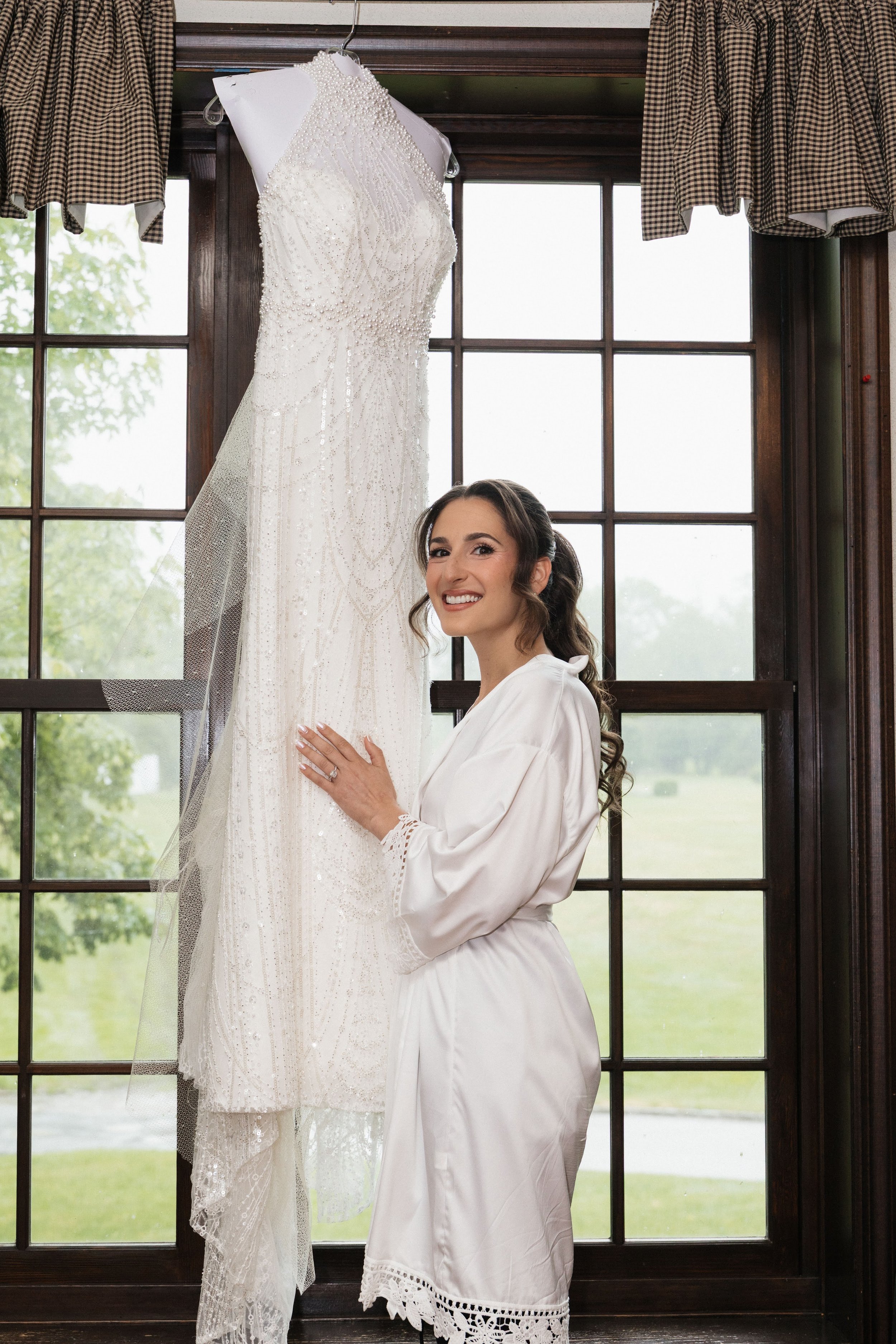 A woman in a white robe is smiling and looking at a white lace wedding dress hanging on a hanger in front of a window with curtains.