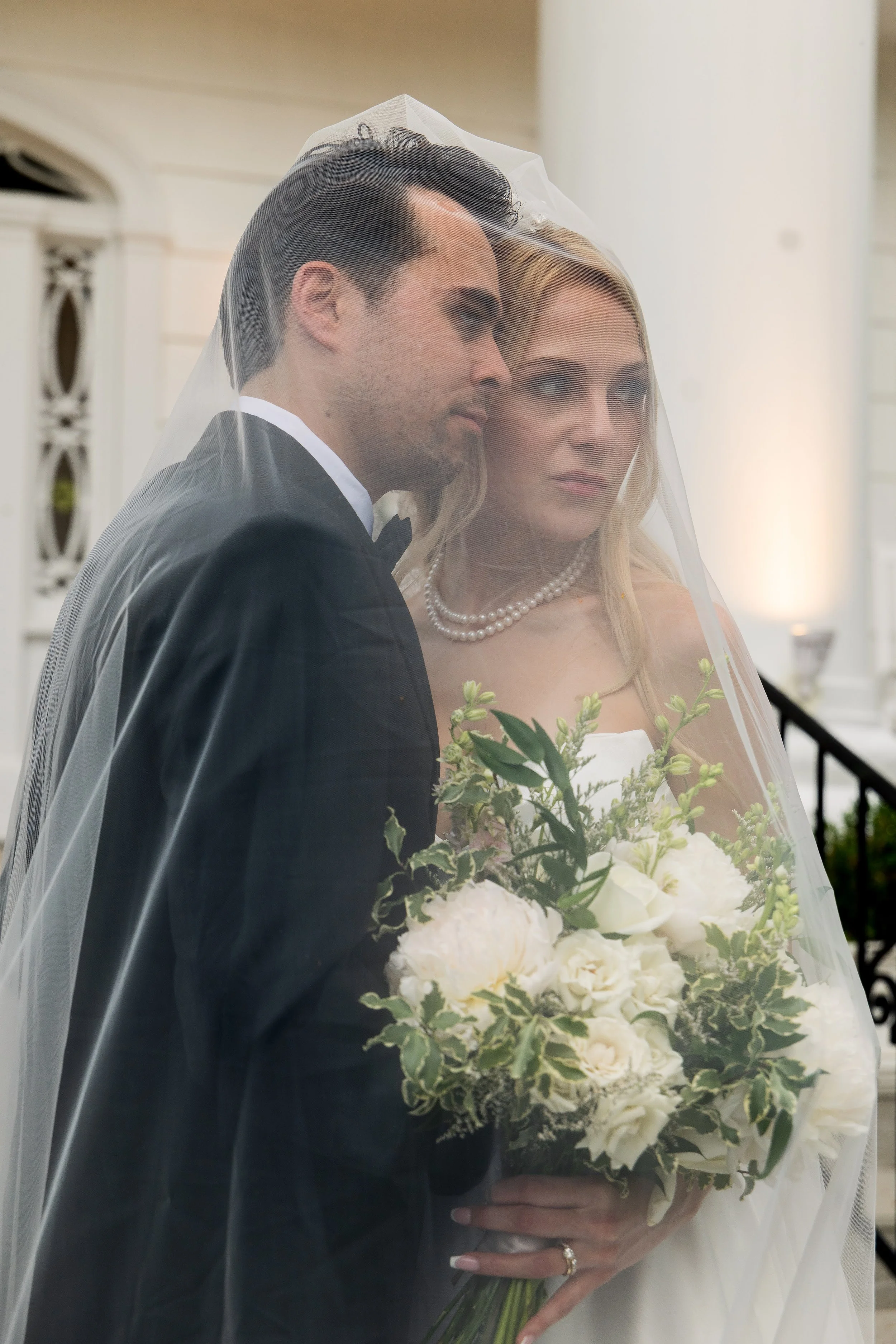 A bride and groom on their wedding day, with the bride holding a white floral bouquet, both covered with a sheer veil. The groom is in a black tuxedo with a bow tie, and the bride wears a pearl necklace.