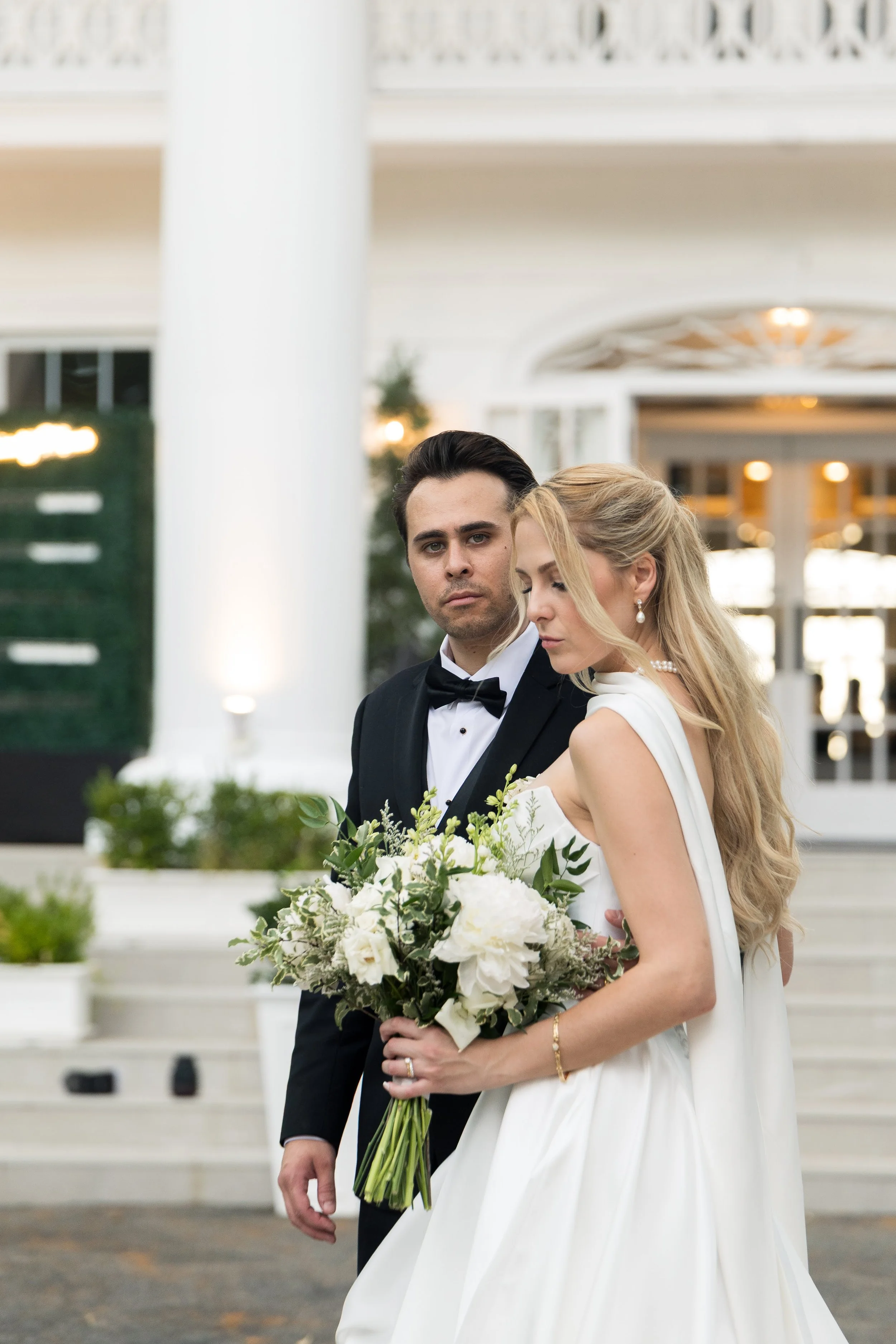 A bride and groom standing together outside, with the bride holding a bouquet of white flowers. The groom is dressed in a tuxedo and the bride in a white wedding gown, both looking solemn. They are in front of a building with white columns and railin