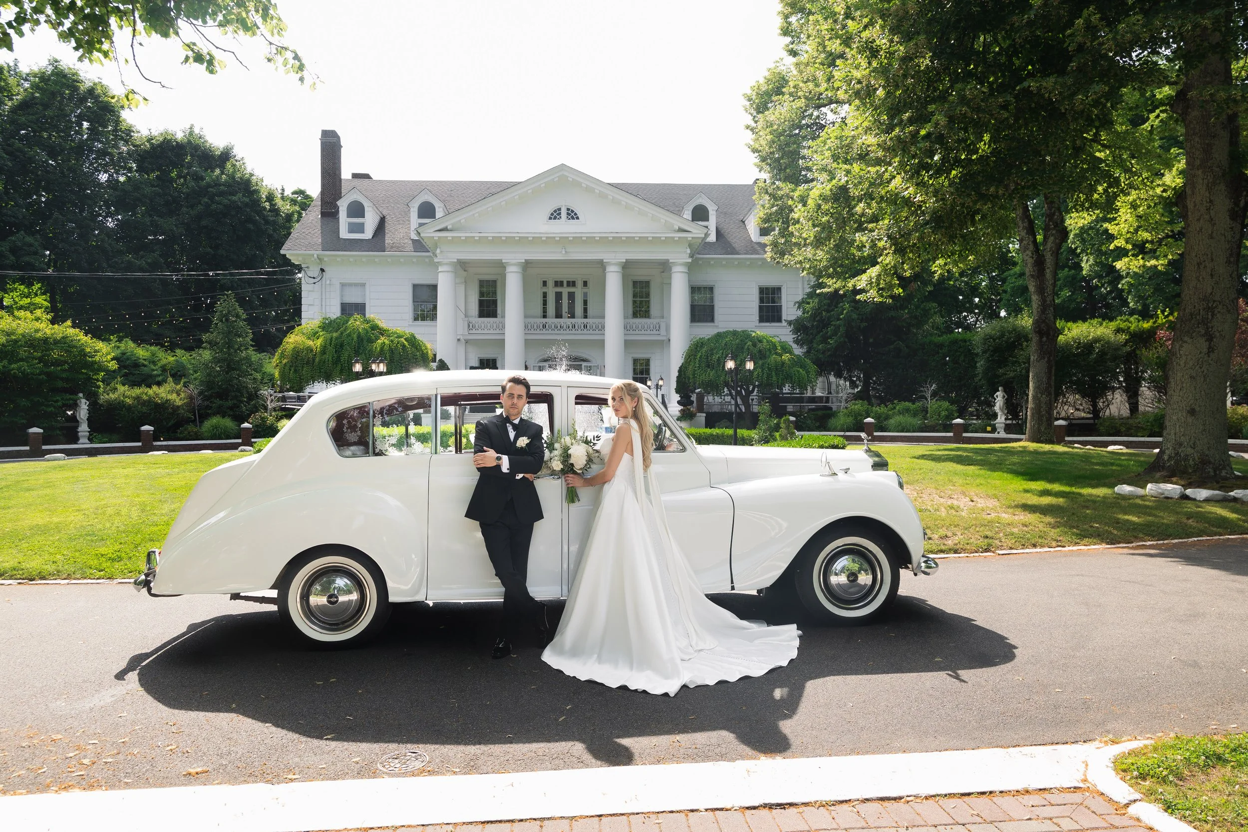 A newlywed couple stands beside a vintage white car in front of a large white house with tall columns. The groom is dressed in a black tuxedo, and the bride in a white wedding gown holding a bouquet. The scene is set outdoors on a sunny day with gree