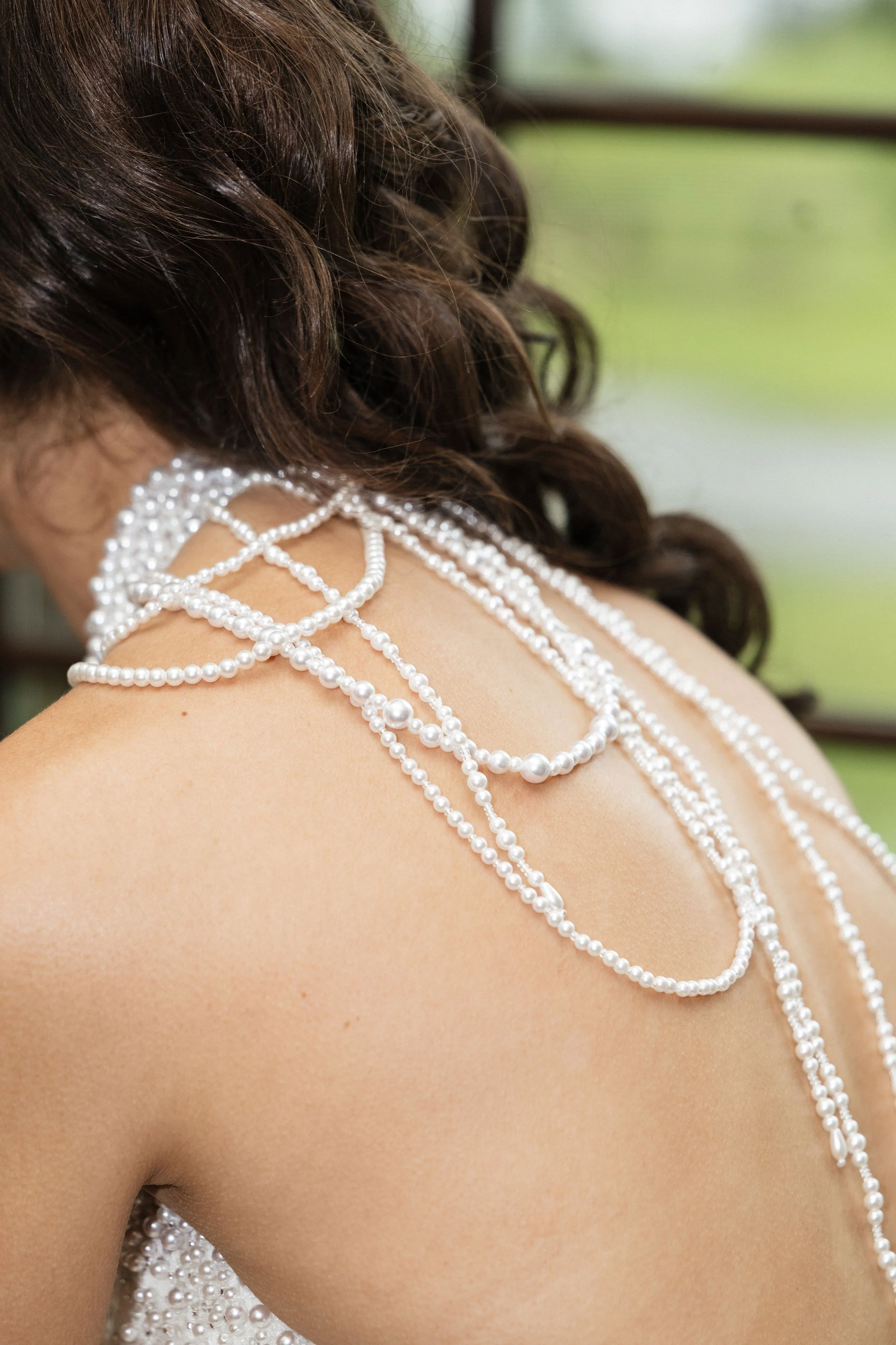 Close-up of a woman with dark, curly hair, wearing multiple strands of white pearls on her shoulder and back, outdoors with a blurred green background.