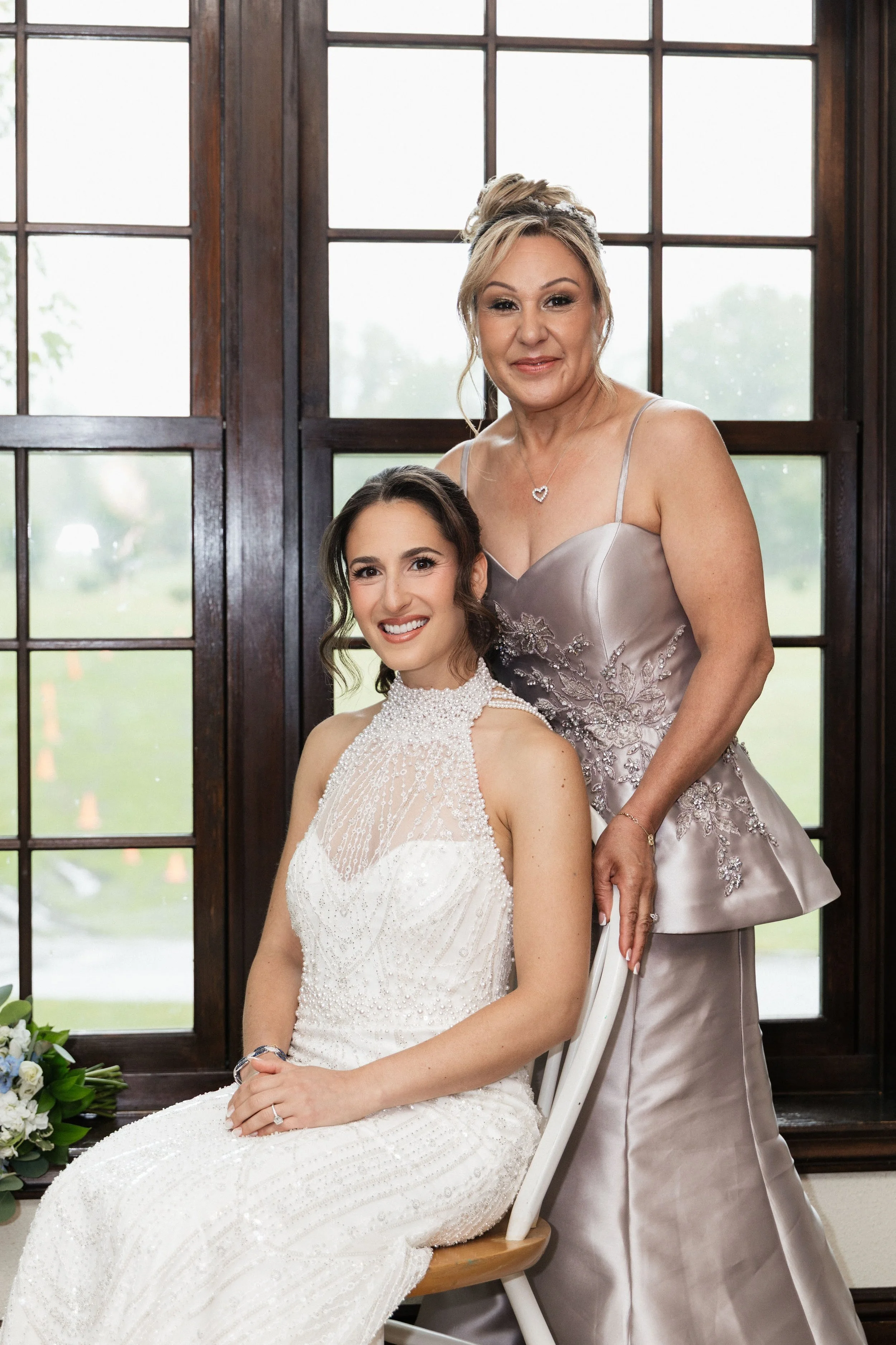 A bride sitting in a chair with a woman standing behind her, both smiling, in front of large windows with outside greenery.