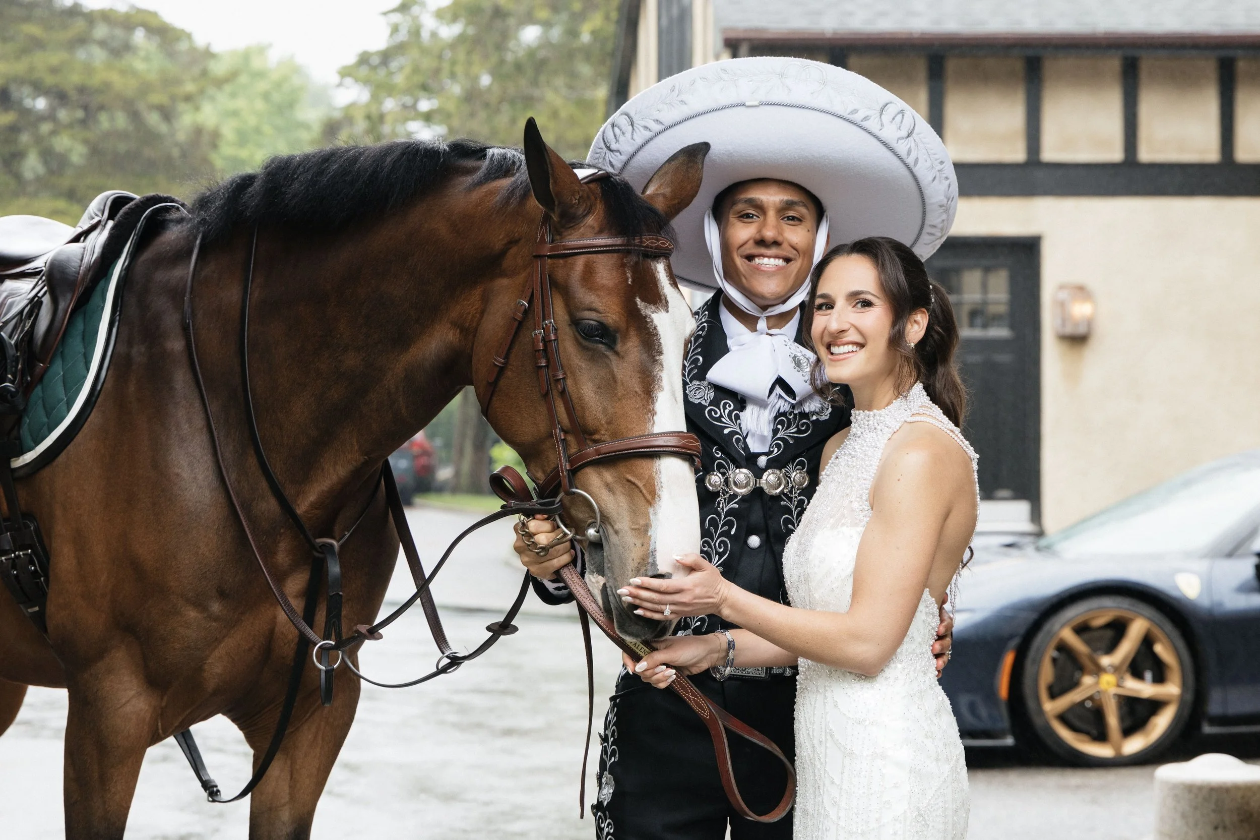 A smiling man and woman dressed in wedding attire standing next to a brown horse with a white stripe on its face, at a rustic outdoor location.
