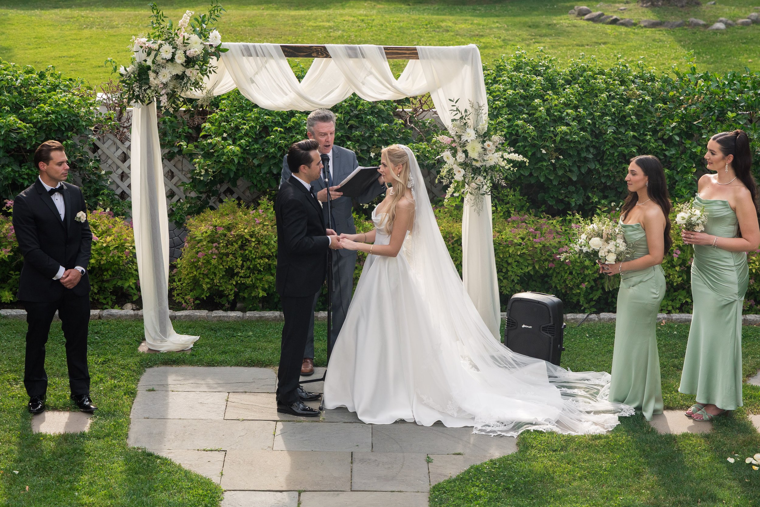 A wedding ceremony outdoors with a bride and groom holding hands, a man officiating, and bridesmaids standing nearby, under a decorated arch with flowers.