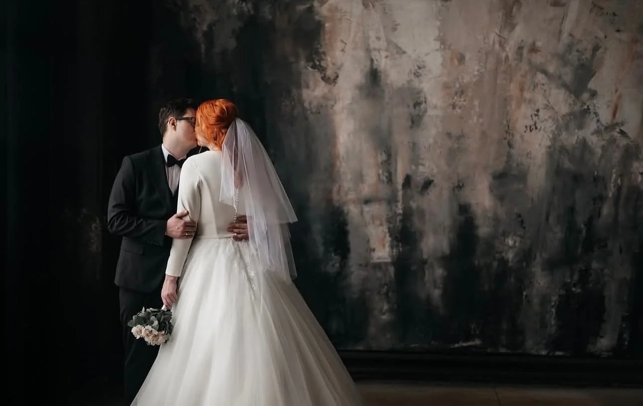A bride and groom kiss in front of a textured dark wall, with the bride holding a bouquet of flowers.