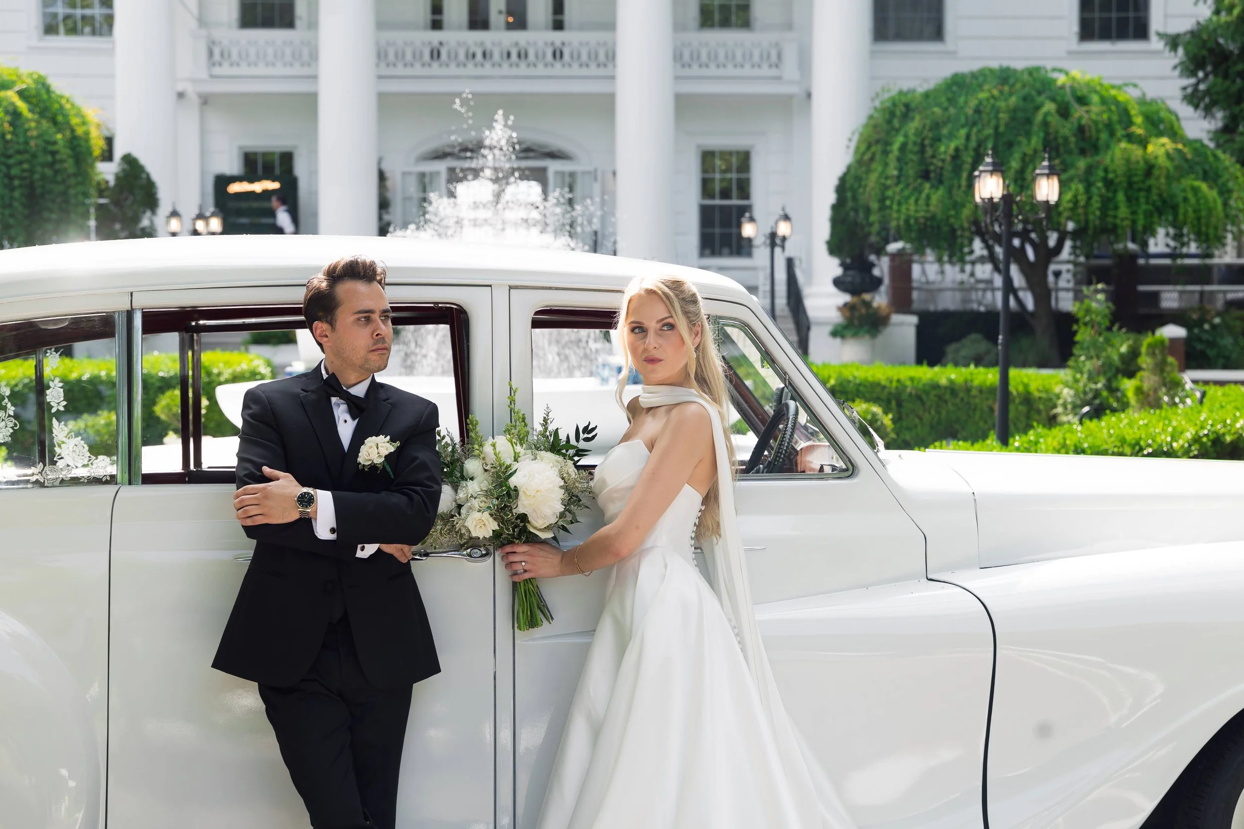 A bride and groom stand beside a vintage white car in front of a grand white building, with the bride holding a bouquet of white flowers and the groom dressed in a tuxedo with a bow tie, both looking in the same direction.