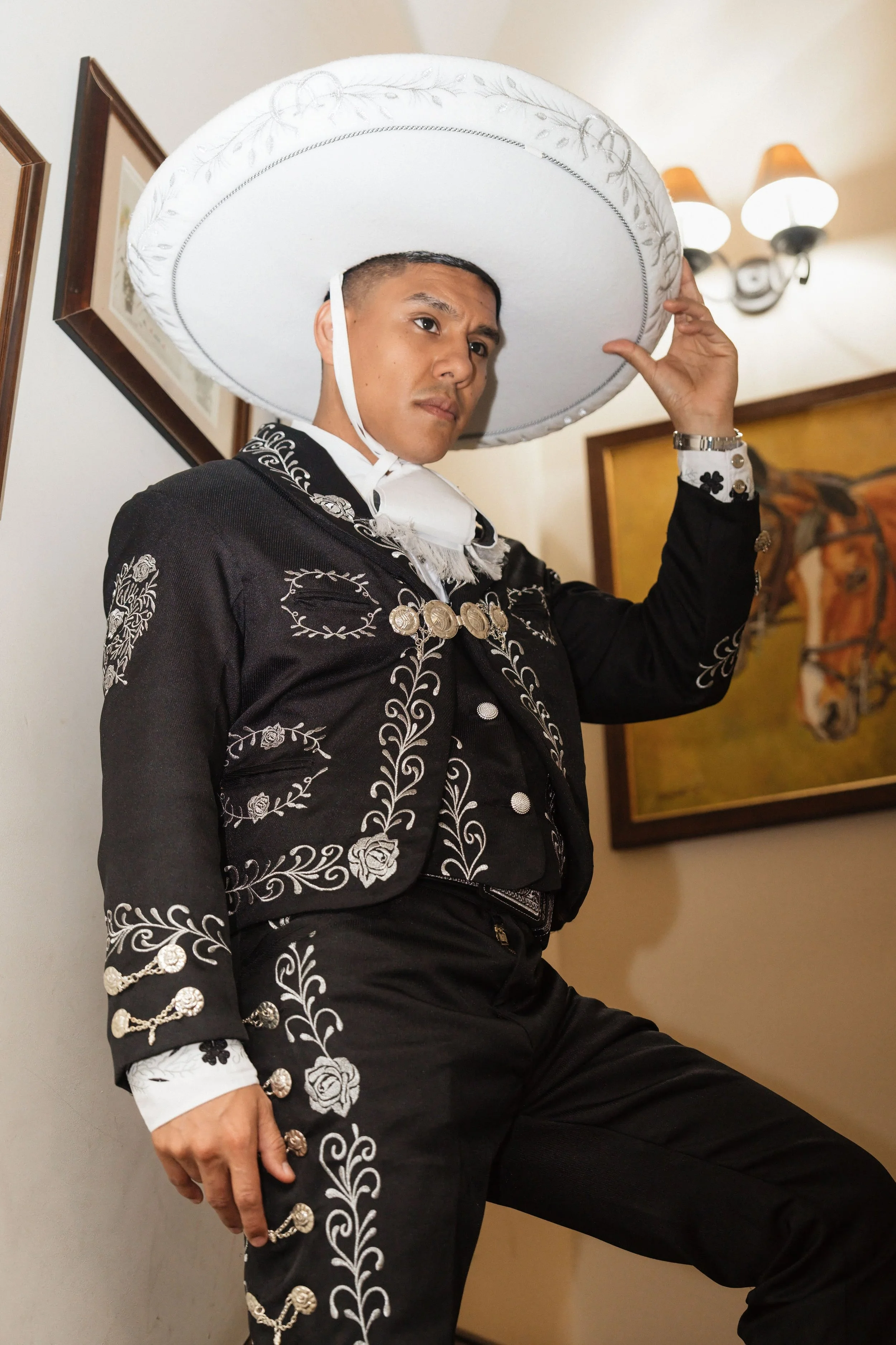 A man wearing a traditional black charro suit with white embroidered floral designs and decorative silver buttons, holding a large white sombrero with silver embroidery, standing indoors with paintings on the wall behind him.