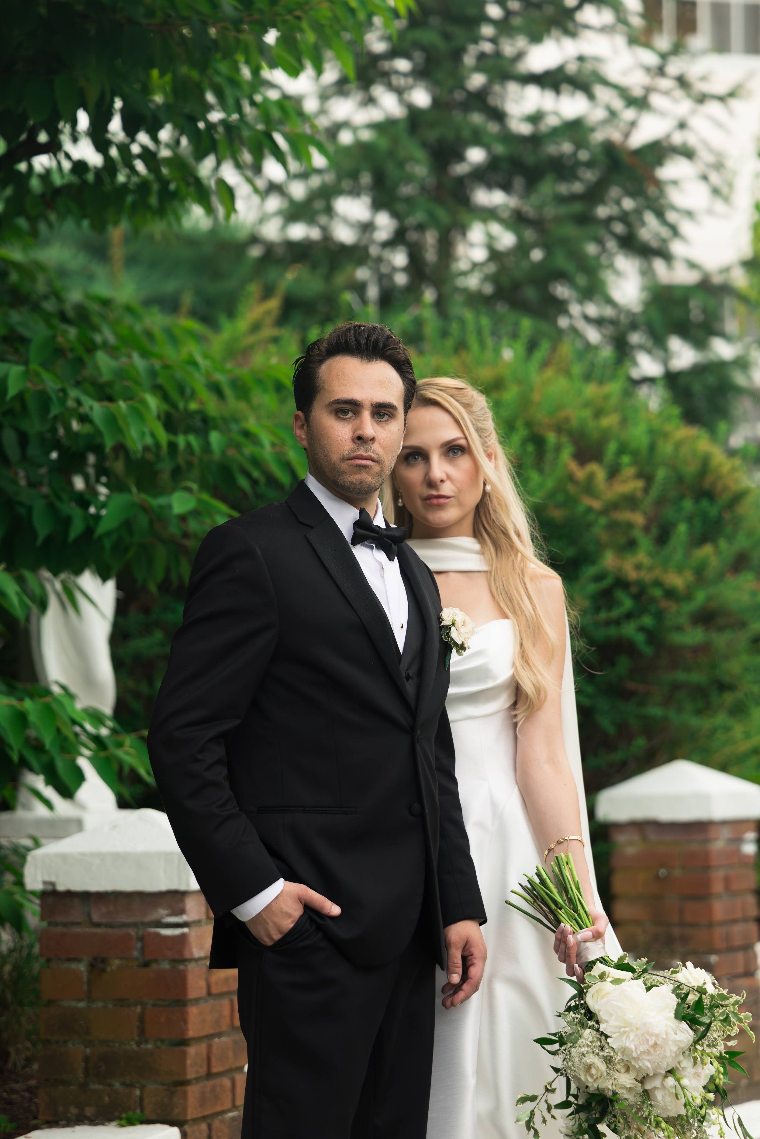 A man in a tuxedo and a woman in a white dress posing outdoors at a wedding with greenery and brick structures in the background.