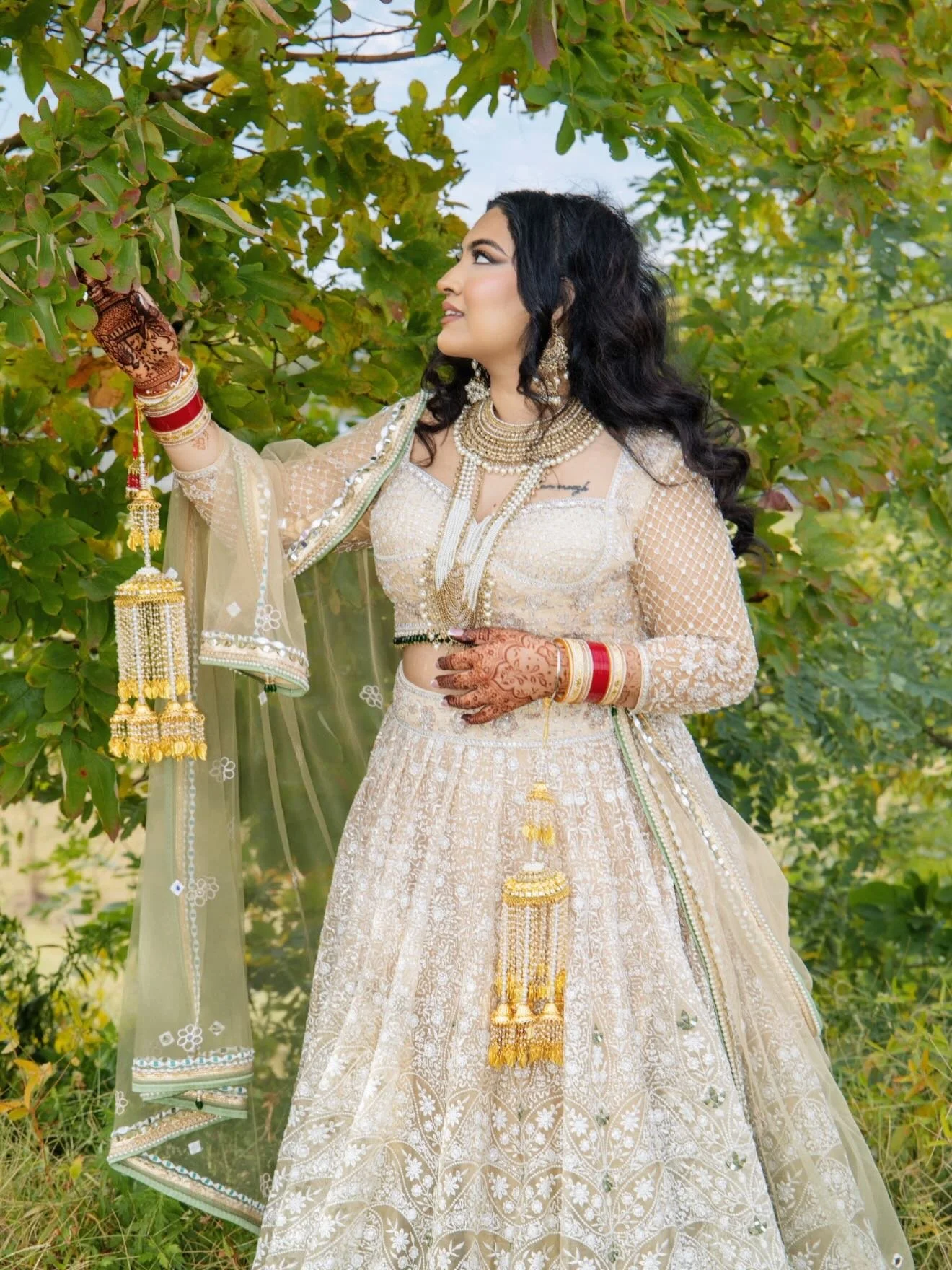 A woman dressed in traditional Indian attire standing outdoors next to green foliage, looking to her left, surrounded by jewelry and detailed embroidery on her cream-colored dress.