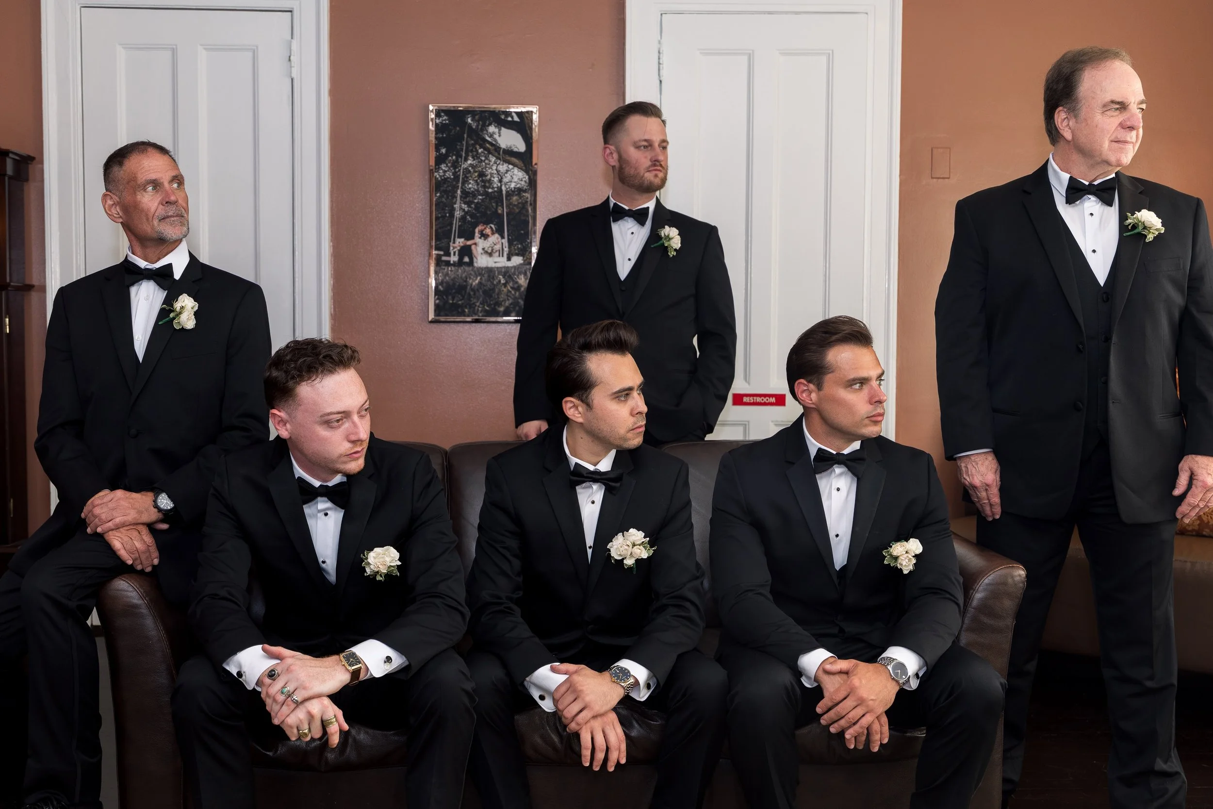 A group of men dressed in black tuxedos with bow ties, seated and standing in a room with brown and white walls, some with white boutonnieres on their lapels, during a formal event or wedding.