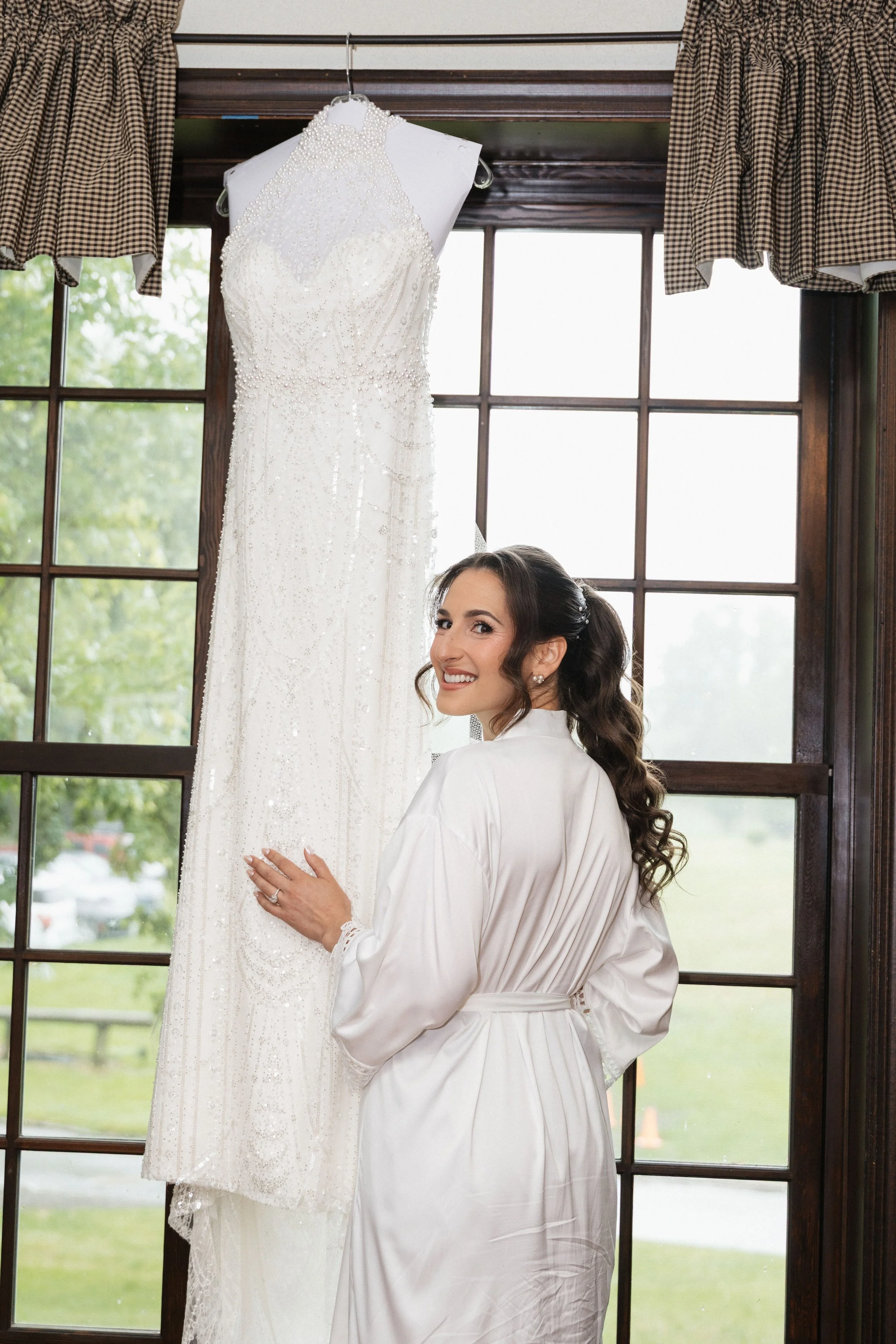A woman in a satin robe standing next to a wedding dress hanging in front of a window with checkered curtains.