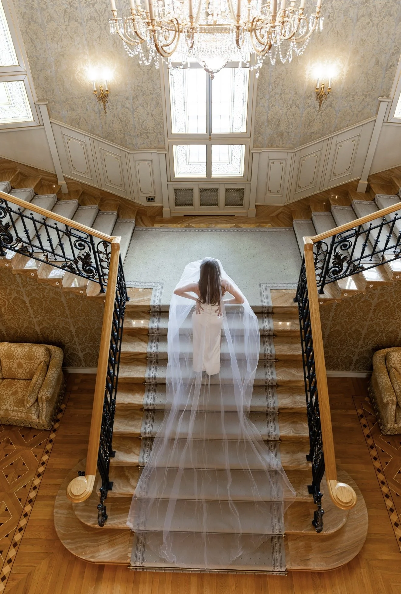 A bride in a wedding gown with a long veil stands on a staircase in an elegant, gold-colored, and floral-patterned room with chandelier lighting.