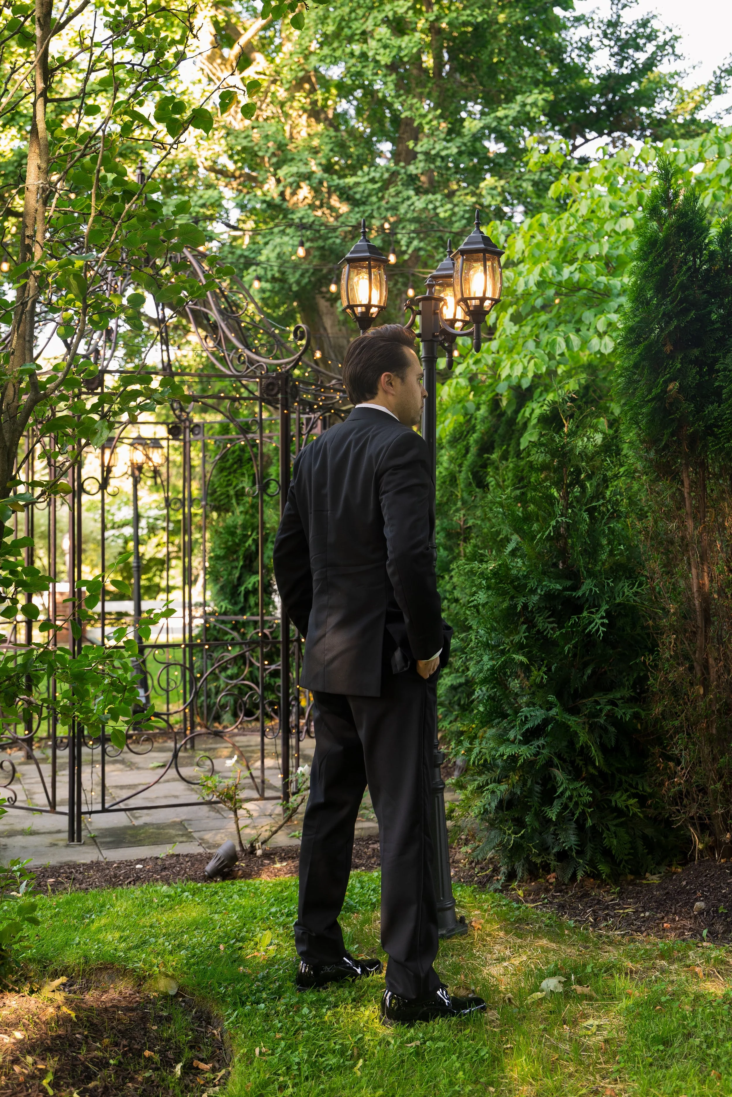 Man in a suit stands outdoors near a lamp post, surrounded by lush green trees and bushes, with decorative wrought iron fencing in the background.