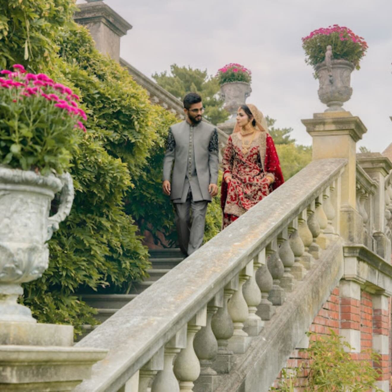 A man and a woman dressed in traditional South Asian attire walking down a stone staircase outdoors, surrounded by lush greenery and flowering plants.