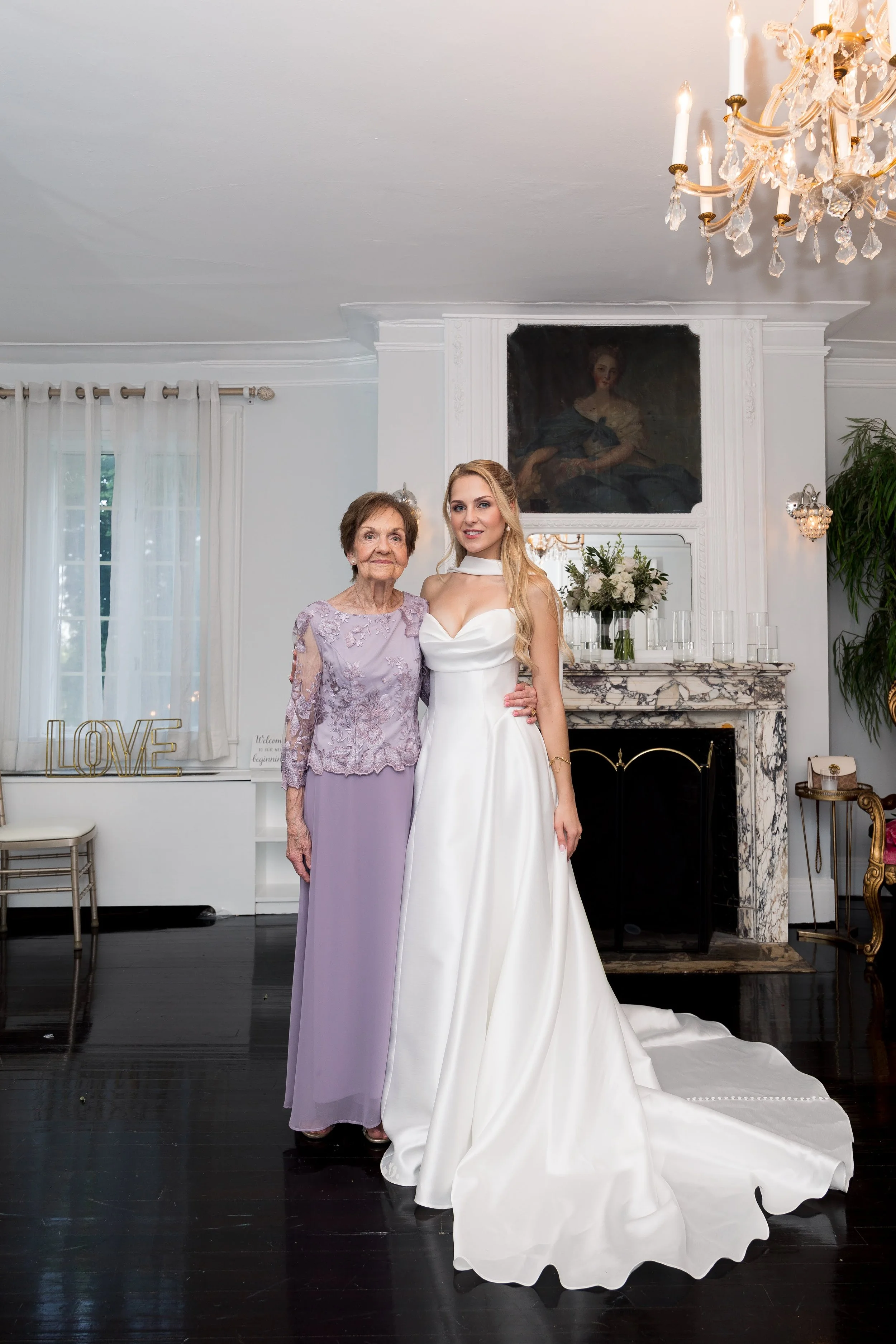 A young woman in a white wedding dress standing next to an elderly woman in a lavender dress in a bright, elegant room with a marble fireplace, a large framed portrait above the fireplace, a crystal chandelier, and white curtains.