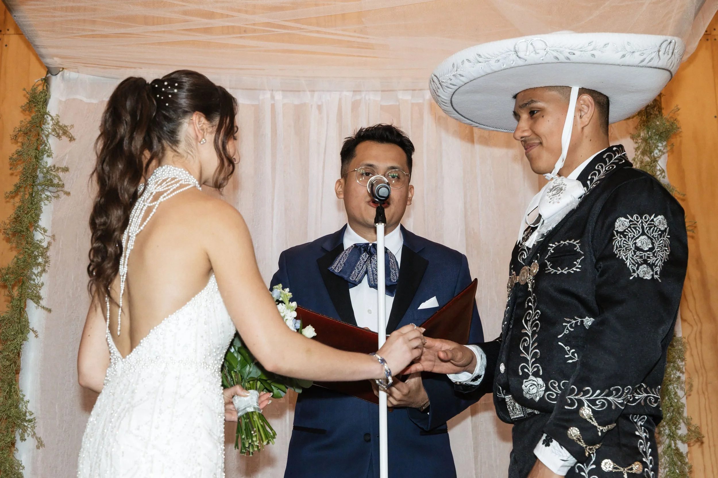 A couple getting married, facing each other and holding hands, with a person officiating in the middle, in a decorated venue with curtains and greenery.