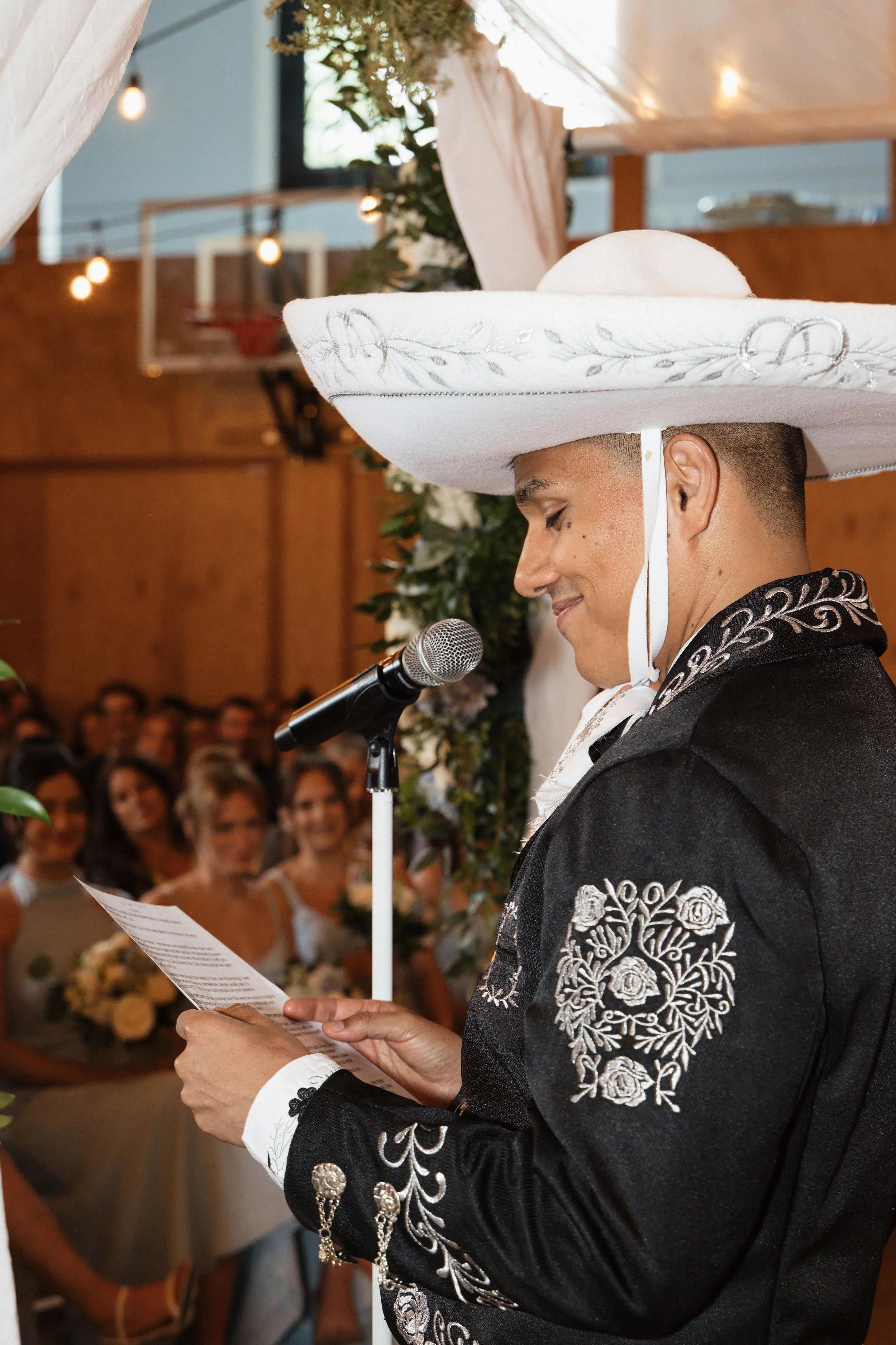 A person wearing a traditional Mexican charro suit reading a speech at a wedding ceremony. The person wears a large white sombrero with intricate embroidery, and the suit features detailed embroidery, including roses on the sleeve. The background sho