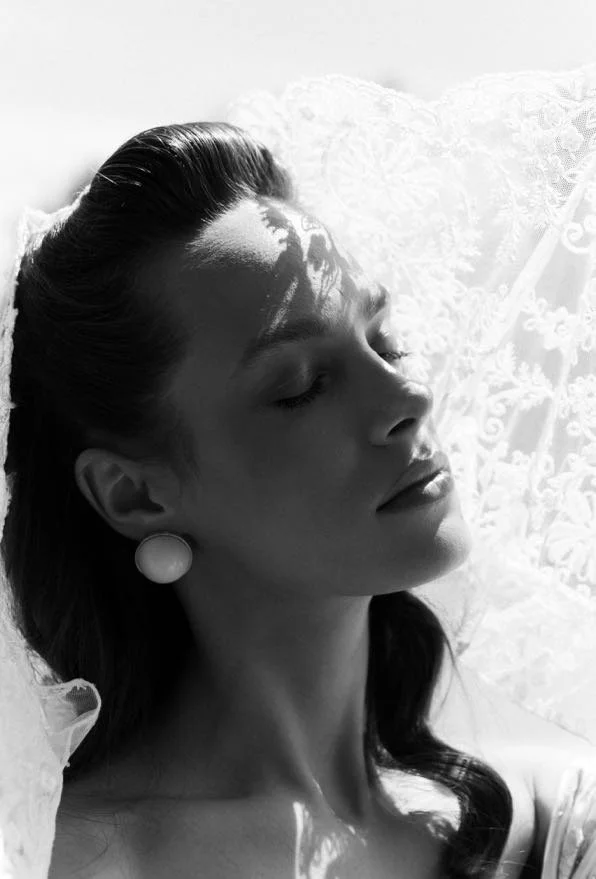 A black and white photograph of a woman with long dark hair, closed eyes, and wearing large round earrings, lying on a textured surface with soft lighting.