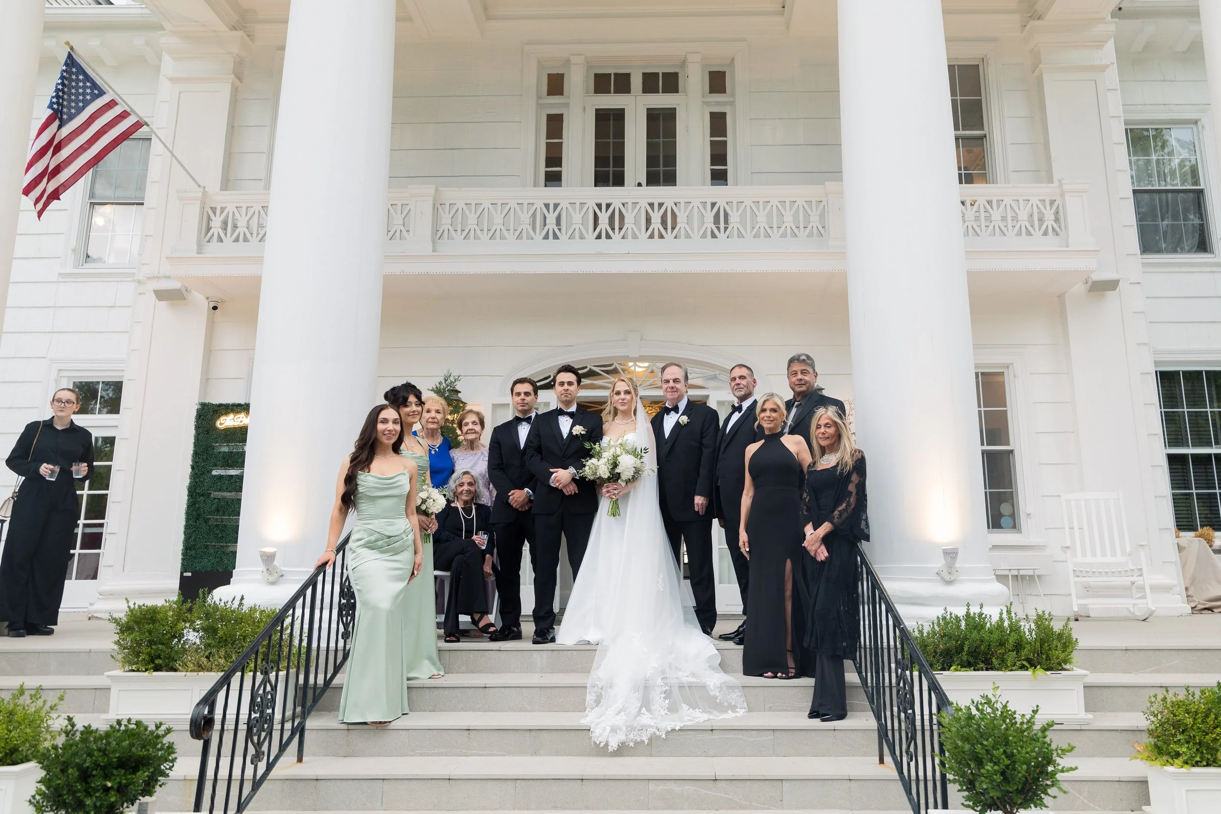 A group of people, including a bride and groom, poses on the steps of a large white house with columns and an American flag, likely celebrating a wedding.