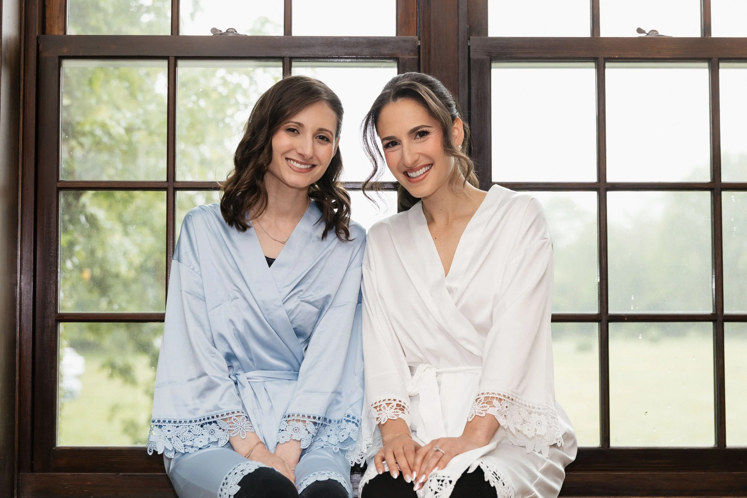 Two women sitting close together by a wooden window, smiling at the camera, one wearing a light blue dress and the other in a white dress with lace details.